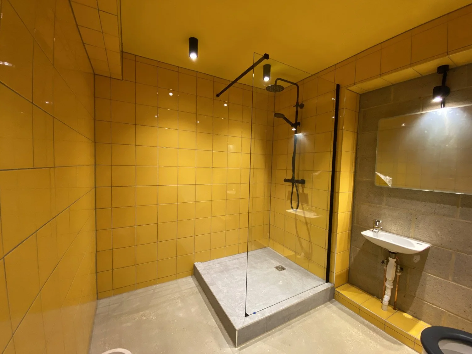 Modern bathroom with yellow tiled walls, a glass shower enclosure, a rain showerhead, a handheld shower, a small wall-mounted sink, a mirror, and ceiling spotlights.