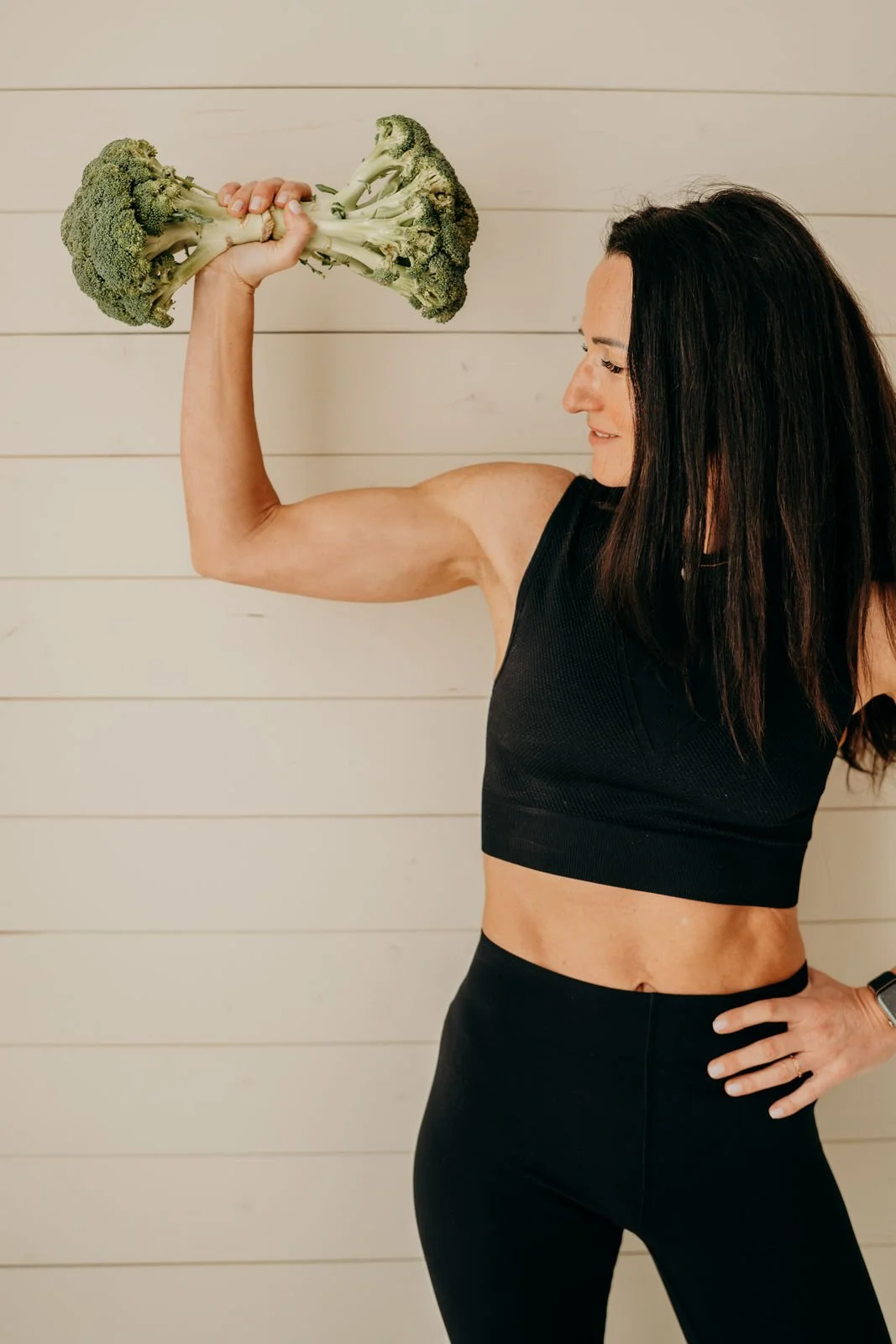 Eine fitnessbegeisterte Frau mit schwarzen Haaren, sportlicher Kleidung, die einen Brokkoli in ihrer Muskel-armhaltung präsentiert.