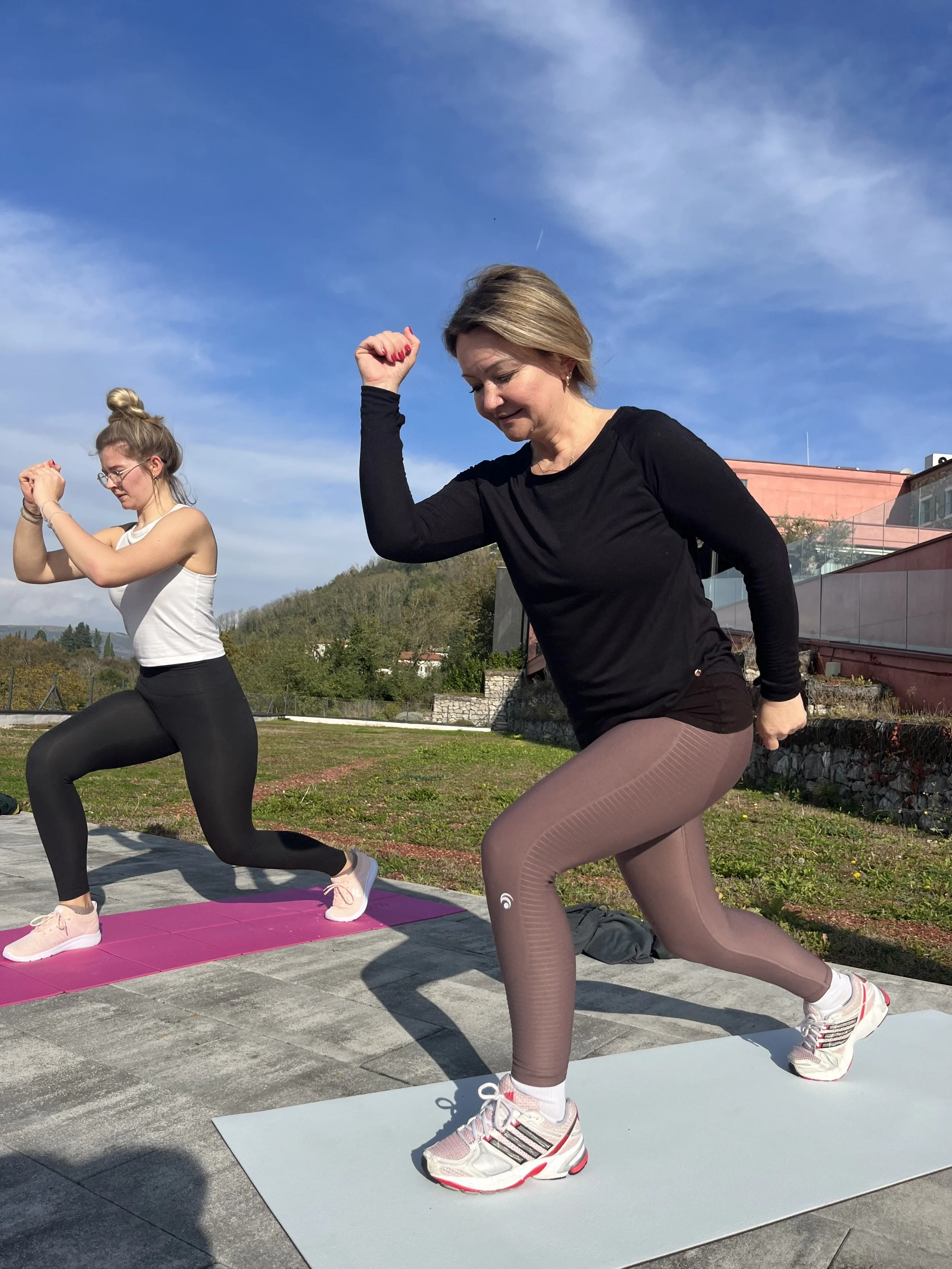 Zwei Frauen machen Yoga im Freien auf einer Wiese, die eine trägt schwarze Sportkleidung, die andere braune Leggings und ein schwarzes Oberteil, beide auf Yogamatten, bei sonnigem Himmel.