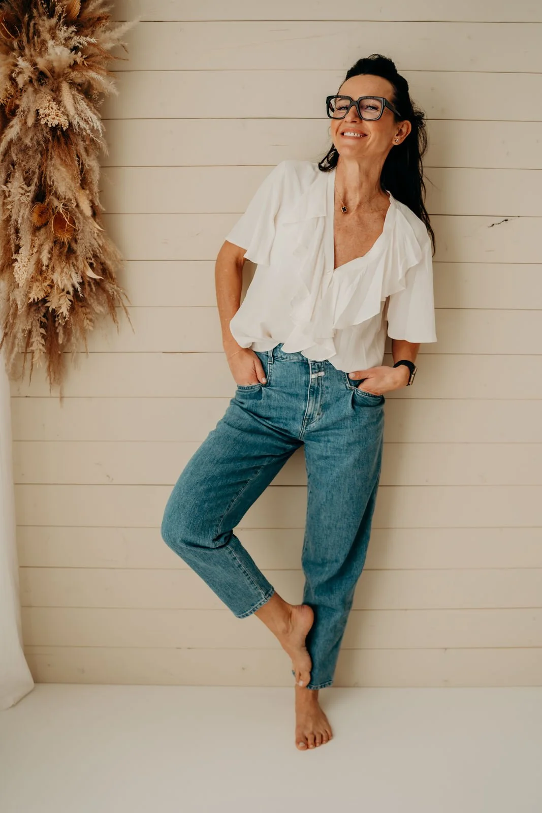 Frau steht lachend vor einer weißen Holz-Wand, trägt eine weiße Bluse und blaue Jeans, ist barfuß, hat dunkle Haare und trägt Brille.