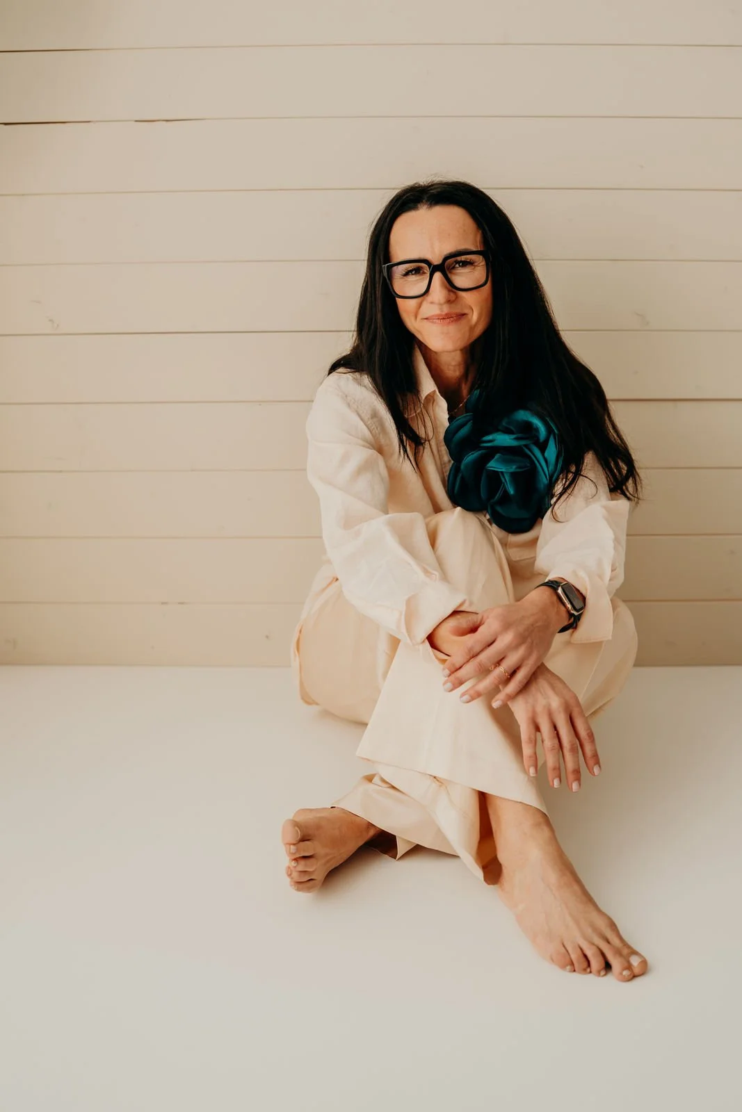 Frau sitzt auf dem Boden, trägt eine cremefarbene Hose und Hemd, trägt eine Smartwatch, hat schwarze Haare, trägt Brille, blickt in die Kamera, vor einem weißen Holzhintergrund.