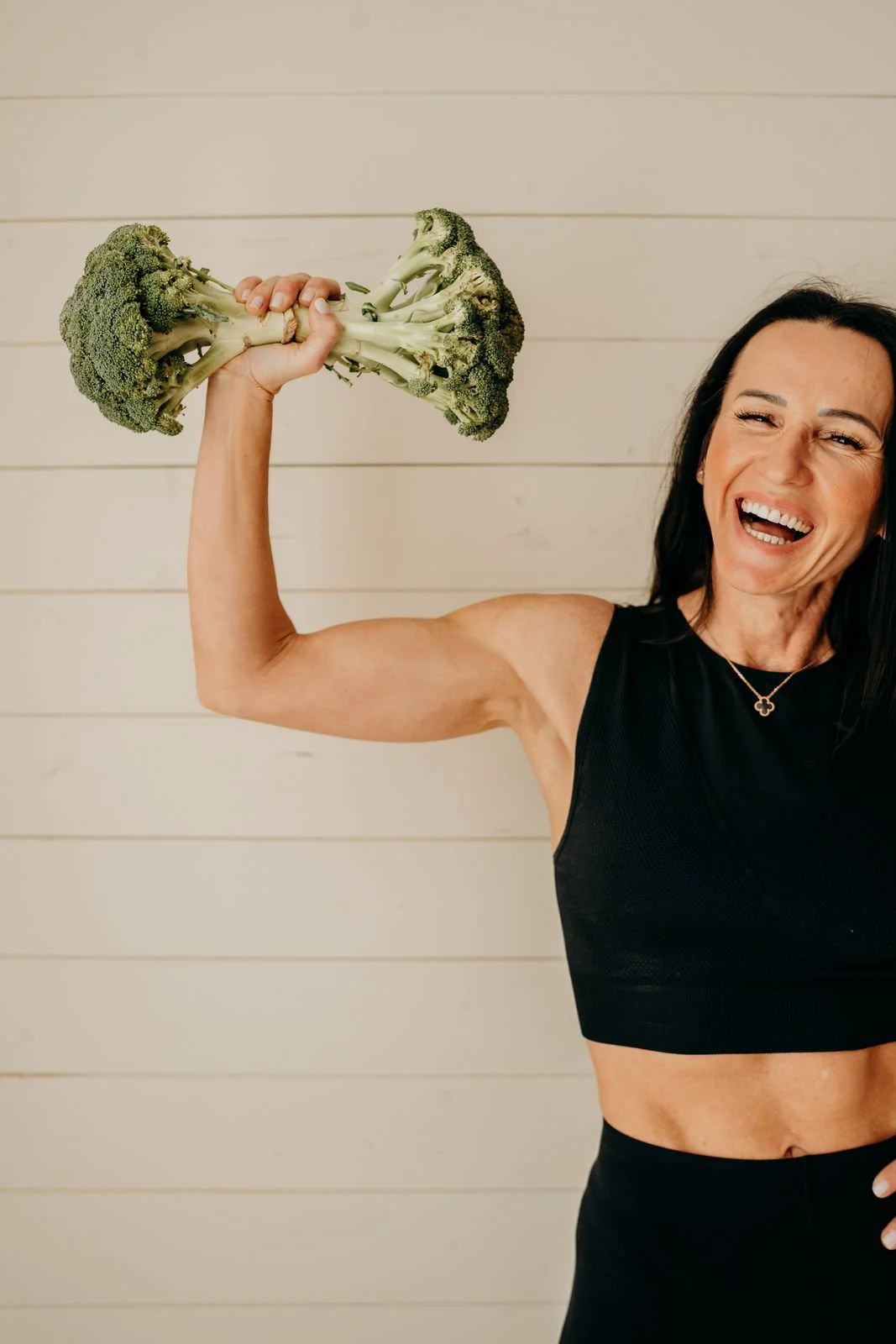 Eine lachende Frau hebt eine Brokkolikopf wie eine Hantel im Fitnessstudio. Sie trägt ein schwarzes Sporttop, die Wand im Hintergrund ist hell und aus Holzpaneelen.