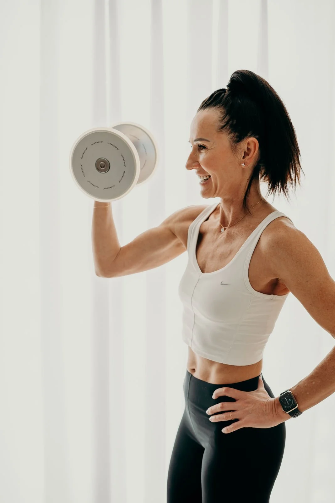 Frau trainiert mit Kurzhantel in einem Fitnessstudio, lächelnd, in Sportkleidung, sie trägt eine Smartwatch.