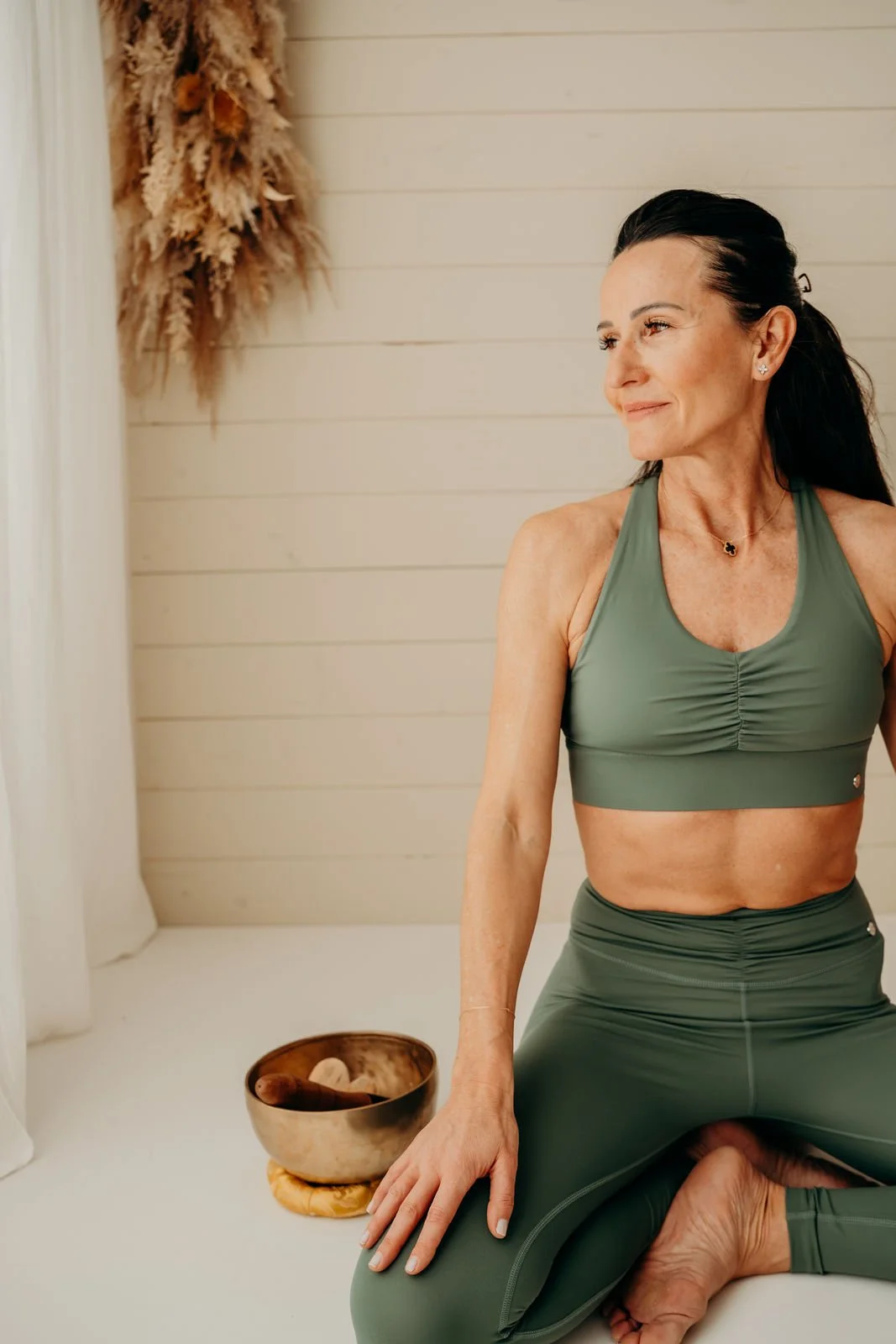 Ein älterer Frau beim Yoga in einem Raum mit weißen Wänden und Trockeblumen im Hintergrund. Sie trägt eine grüne Sport-BH und Leggings, sitzt in einer sitzenden Yoga-Position, neben einer kleinen Schale mit Räucherstäbchen oder anderen Gegenständen.