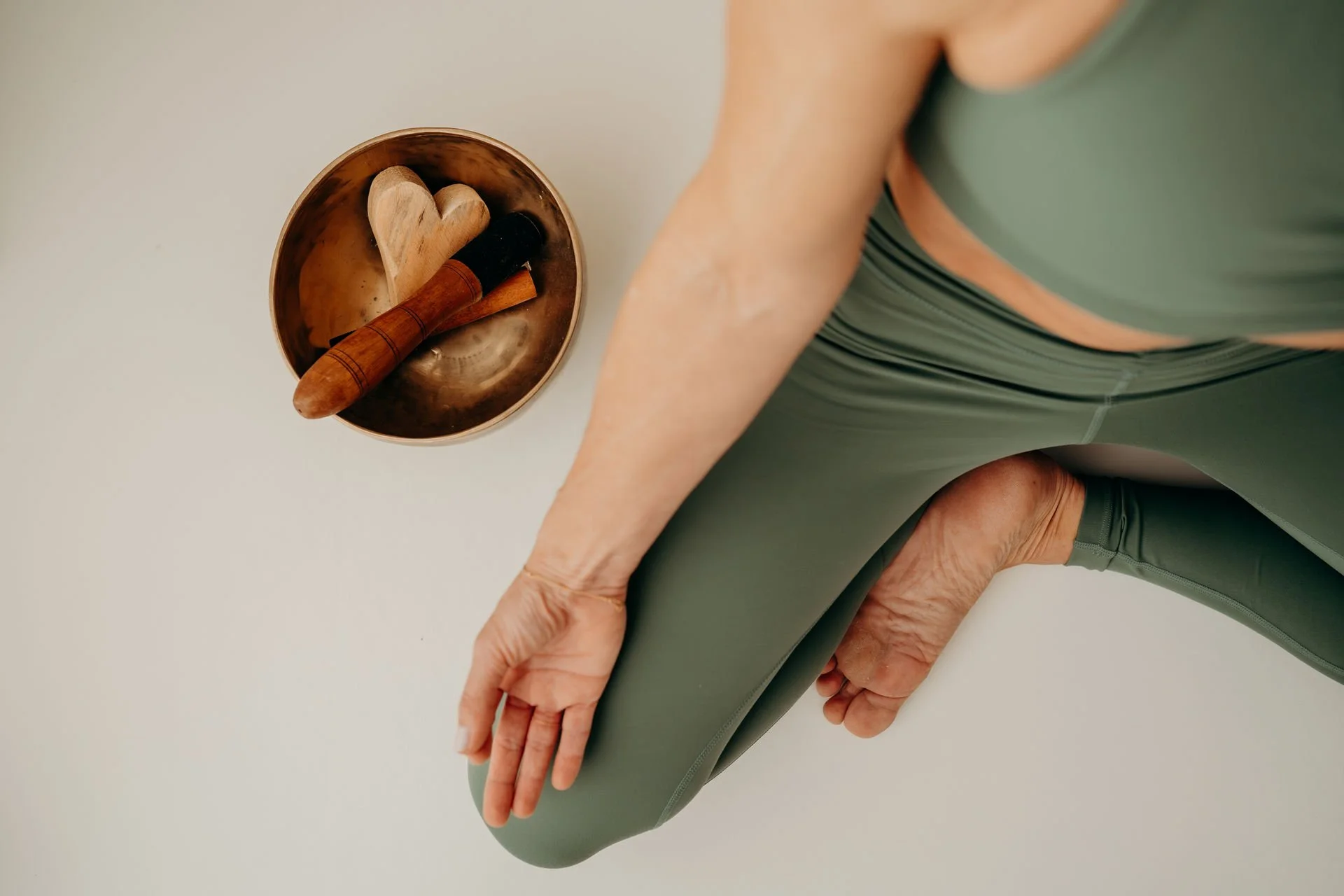 Eine Person in grüner Sportbekleidung führt eine Yoga-Übung durch, während eine Gong-Schale und ein Holzherz auf dem Boden liegen.