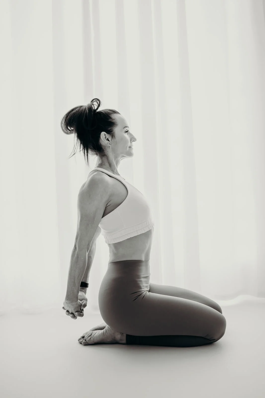 Frau macht Yoga, sitzend auf den geschlossenen Knien und den Armen hinter dem Rücken ausgestreckt, auf einem weißen Boden vor einem weißen Hintergrund.