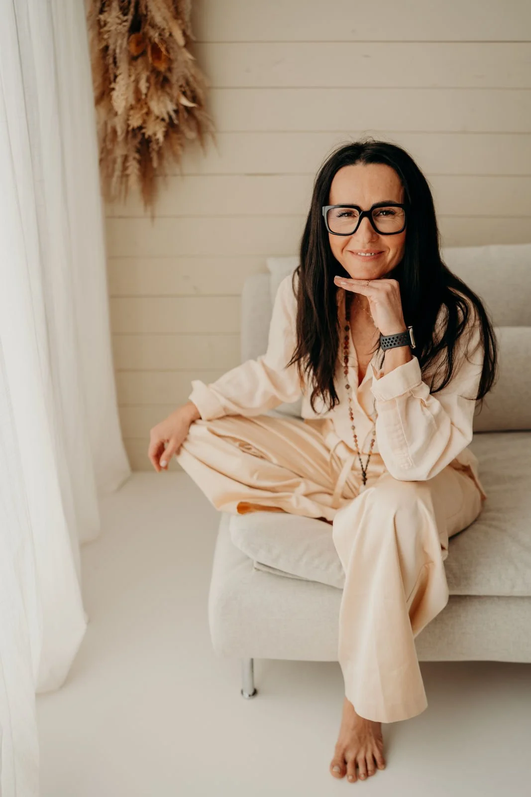 Eine Frau mit schwarzen Haaren und Brille sitzt entspannt auf einer Couch, trägt eine lockere, cremefarbene Kleidung, lächelt und hat eine Hand unter dem Kinn. Im Hintergrund sind weiße Wände und eine Trockenblume an der Wand zu sehen.