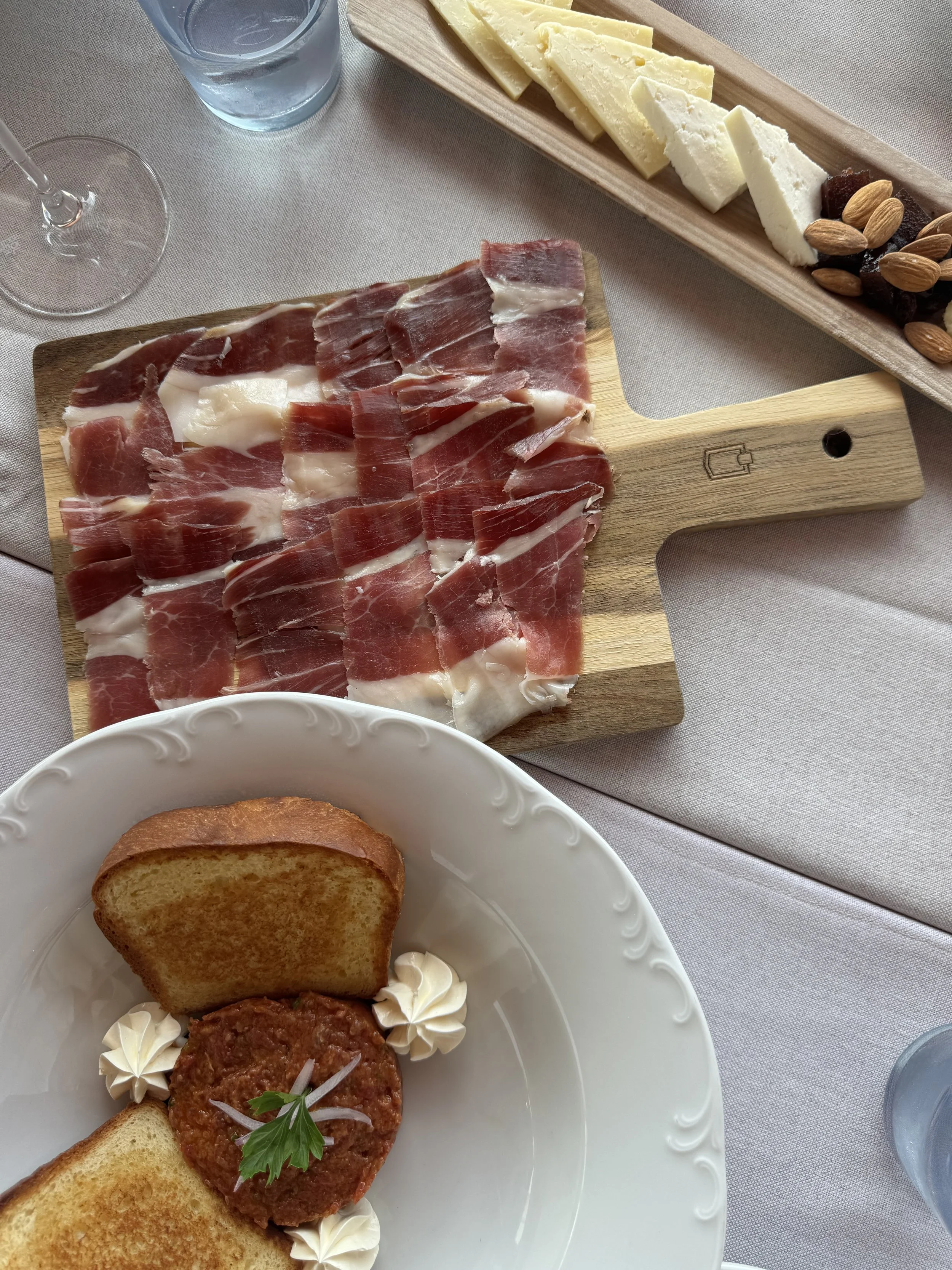 Blick auf eine Käseplatte mit mehreren Käsesorten, einer Platte mit rohem Schinken, einer Scheibe Toastbrot, einer Mahlzeit mit Hackfleischkuchen, einer Scheibe Toast und Tomatensalat, sowie einem Glas Wasser.