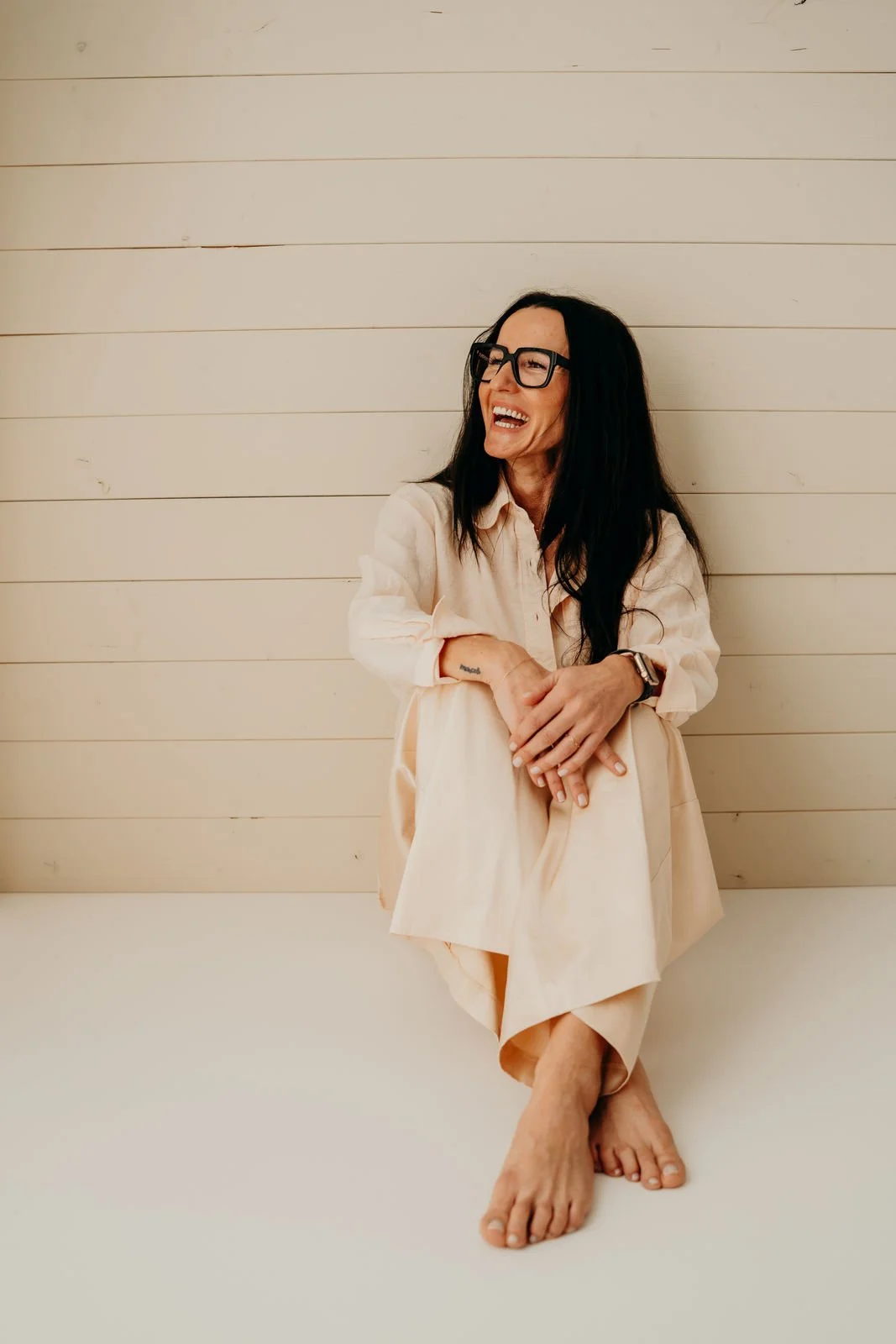 Frau sitzt lachend vor weißer Wand, trägt Brille, helles lockeres Outfit und glattes schwarze Haare, barfuß.