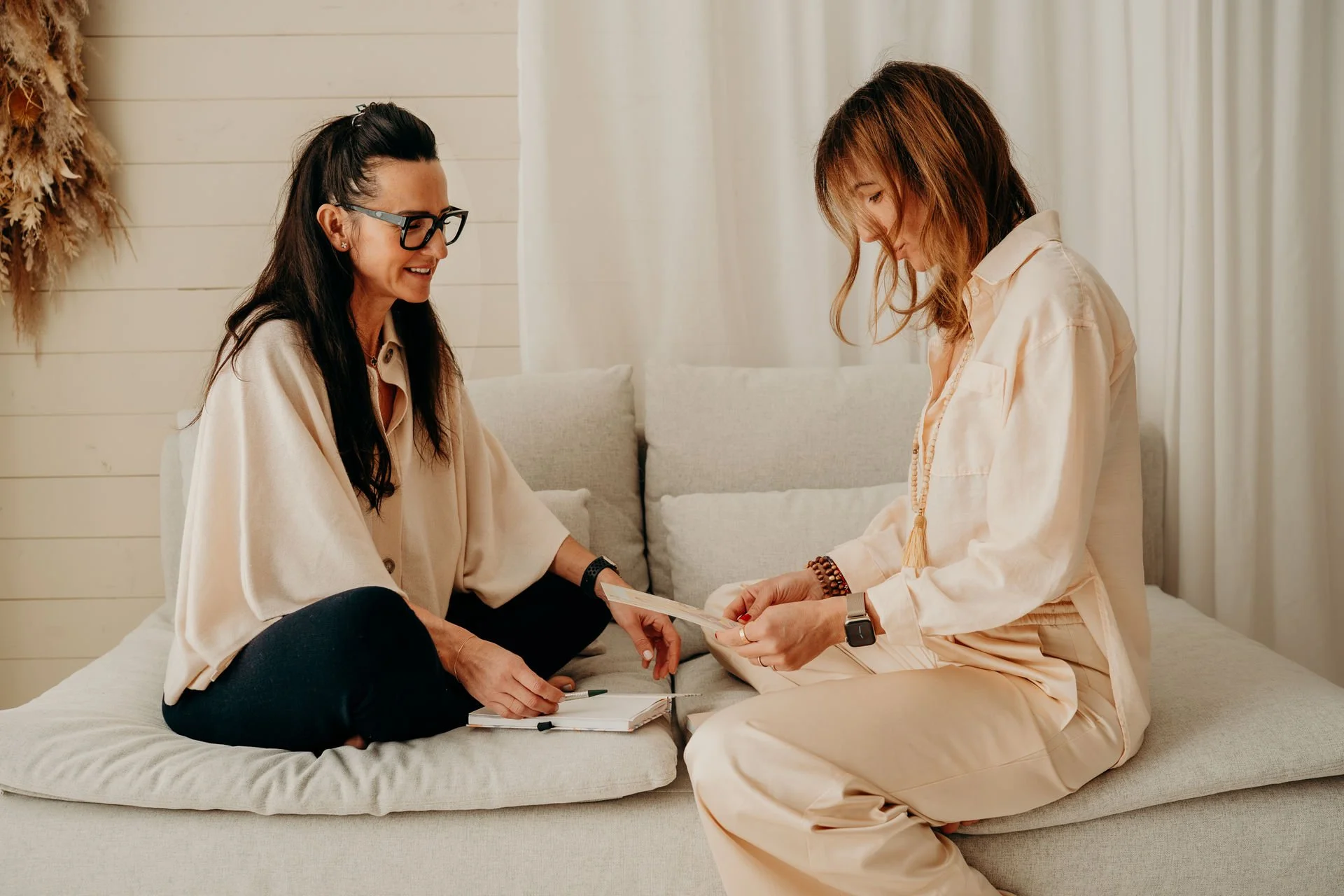 Zwei Frauen sitzen auf einem Sofa, eine Frau mit langen dunklen Haaren und Brille, die andere mit mittellangen braunen Haaren, die ein Journal hält.