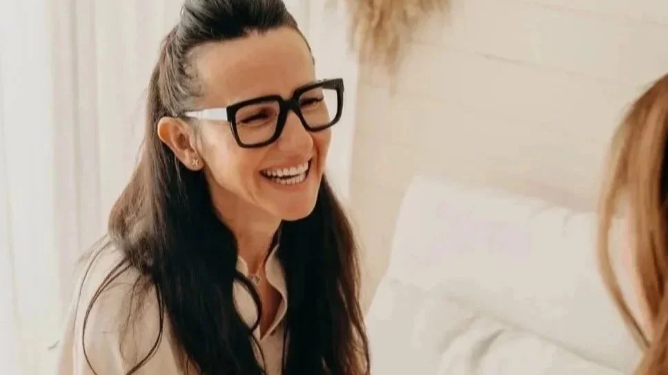 Eine lachende Frau mit dunklem Haar und Brille sitzt auf einem Bett in einem hellen Raum.