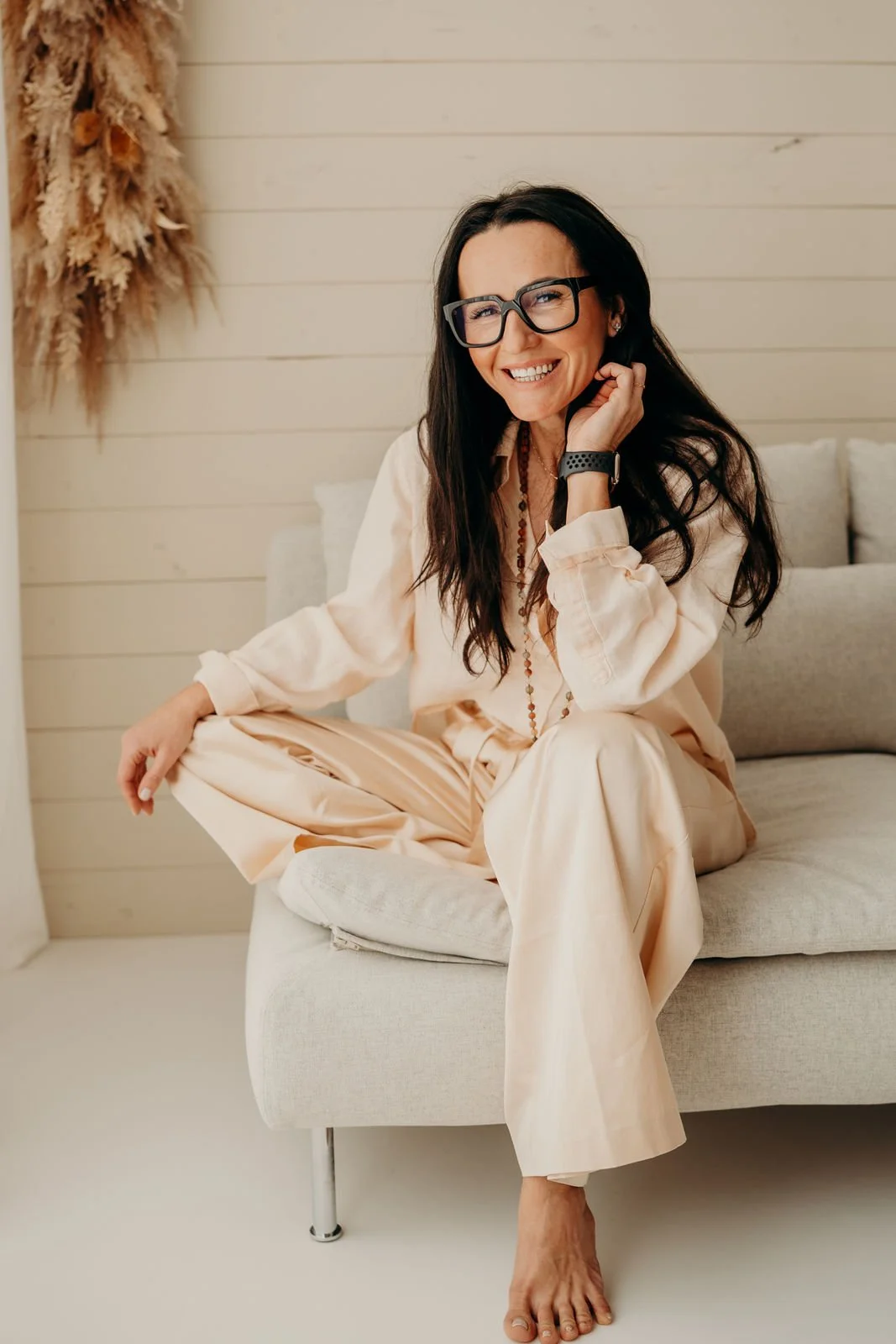 Frau sitzt auf einem Sofa, lächelnd, trägt Brille, dunkle Haare, beige Kleidung, im Hintergrund eine Wand mit Dekor-Trockeblumen.