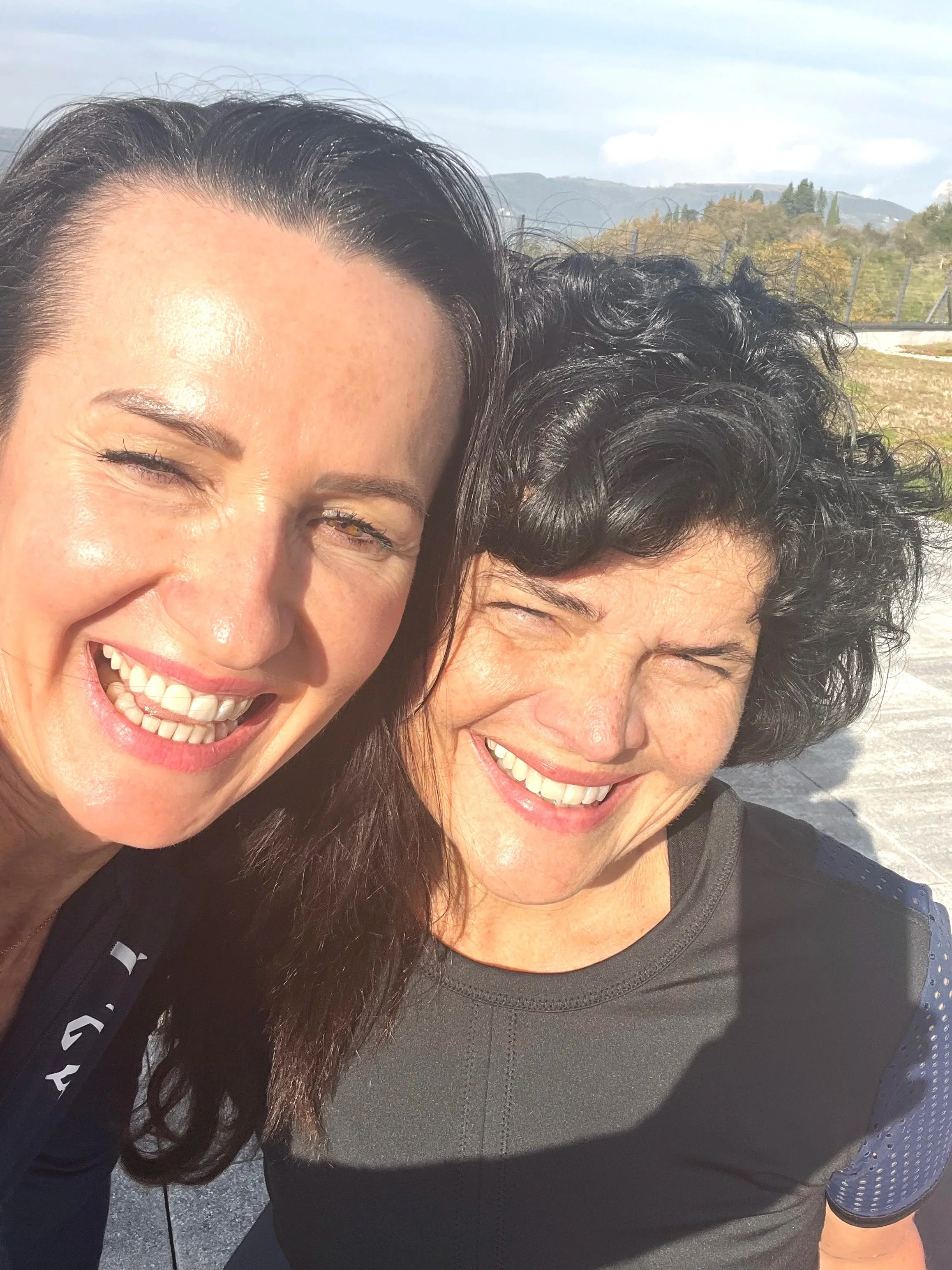 Zwei lachende Frauen beim Outdoor-Selfie, im Hintergrund bergiges Gelände und bewölkter Himmel.