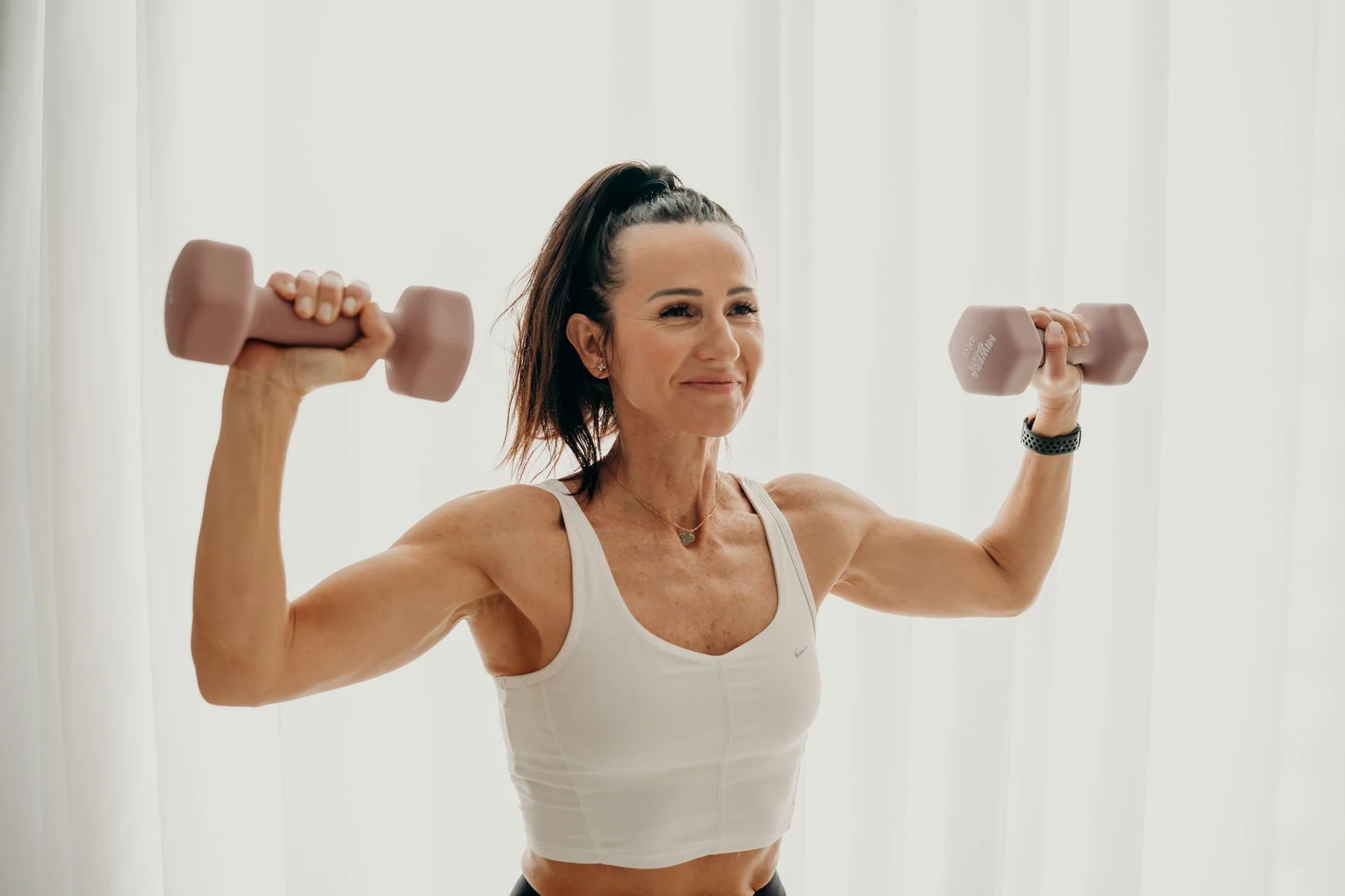 Frau mittleren Alters beim Krafttraining mit Hanteln in einem hellen Raum