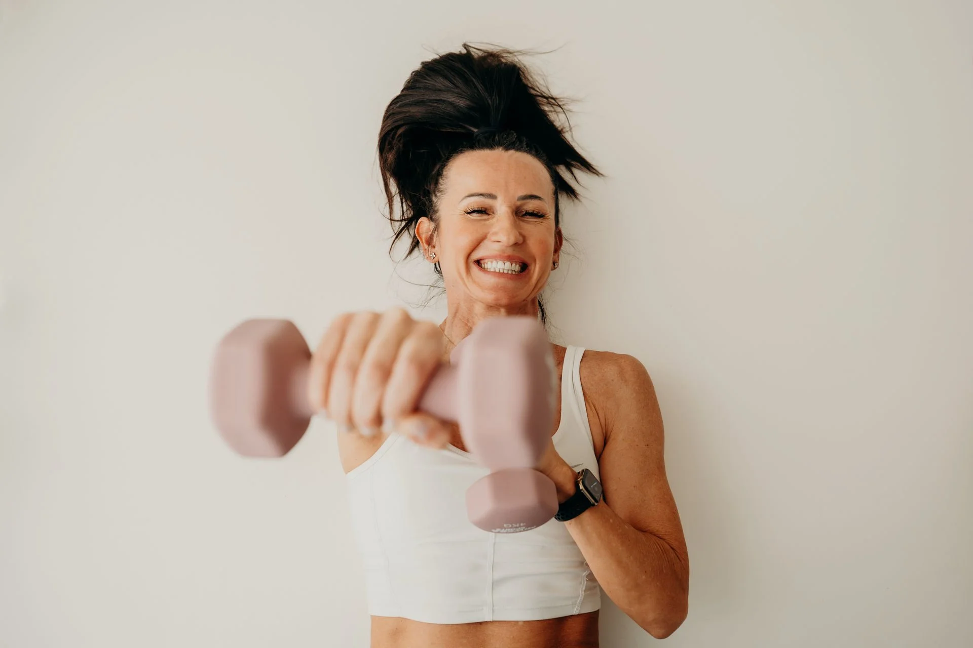 Frau trainiert mit einem pinkfarbenen Hantel, zeigt ein breites Lächeln und trägt Sportbekleidung sowie eine Smartwatch.