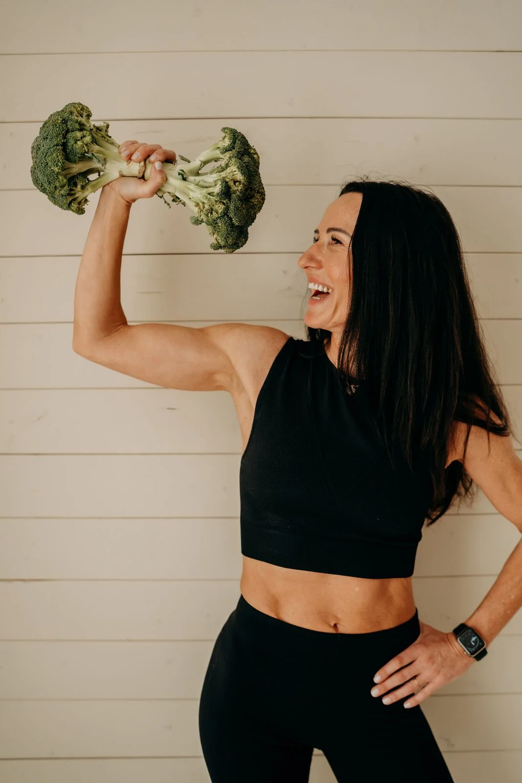 Eine lachende Frau in Sportkleidung, die einen Brokkoli in die Luft hebt wie eine große Hantel.