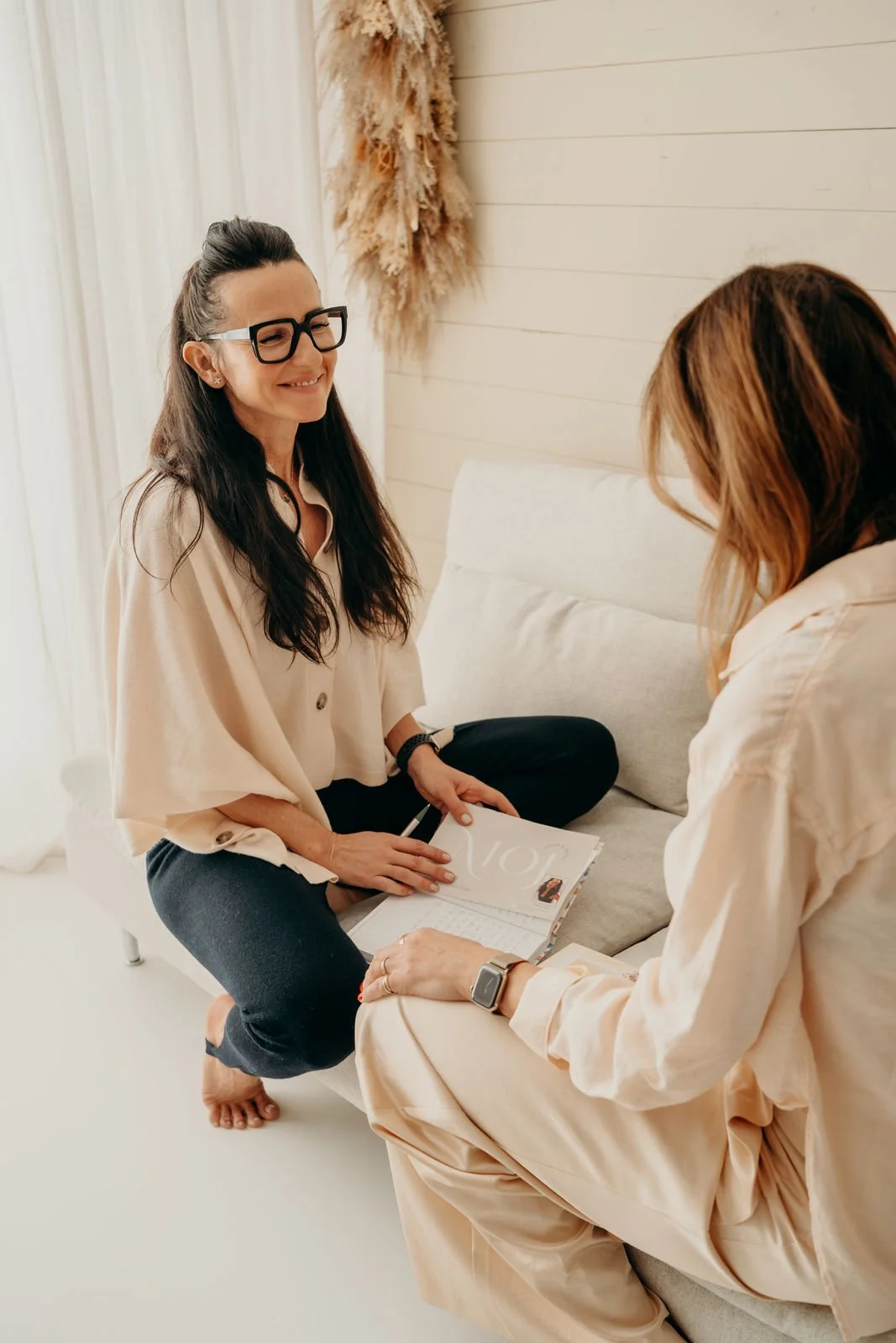 Zwei Frauen sitzen auf einem Sofa, eine lächelt und hat ein offenes Buch oder Magazin, die andere zeigt auf etwas im Buch und trägt eine Smartwatch.