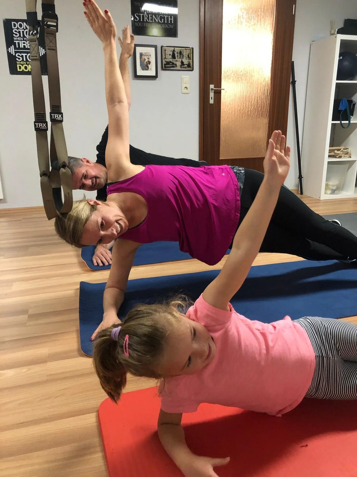 Eine Familie (Vater, Mutter, Kind), die auf Yoga-Matten in einem Fitnessraum gemeinsam trainieren, auf der Seite liegend im Seitenstütz, sie sehen sehr motiviert aus und lächeln