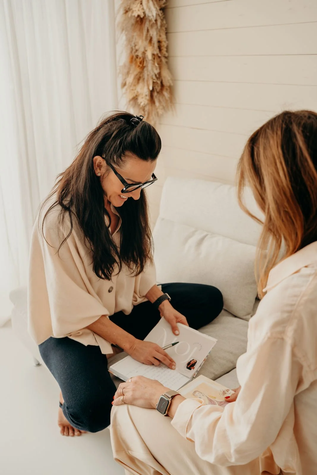 Zwei Frauen sitzen auf einem Sofa, eine zeigt auf ein Notizbuch, die andere schaut zu, in einem hellen Raum.
