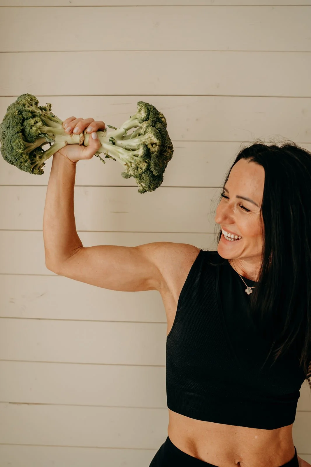 Frau in sportlicher Kleidung hält eine Brokkolipflanze und posiert wie Bodybuilder, lächelt freundlich.
