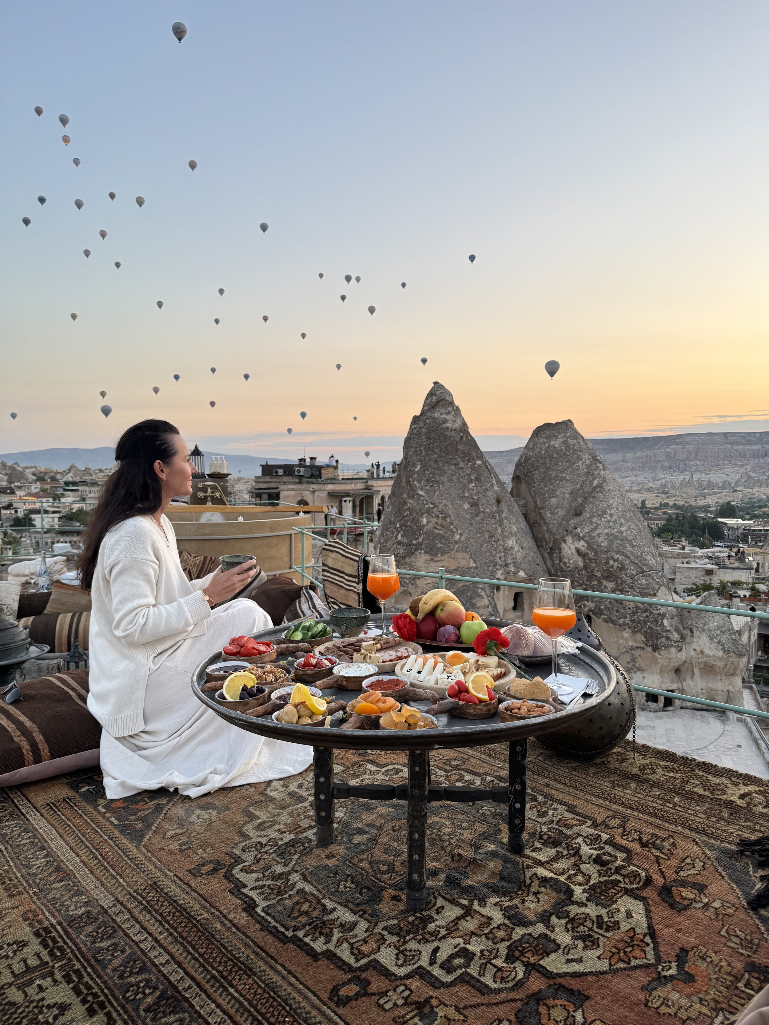Eine Frau sitzt auf einem Teppich auf einem Balkon mit Blick auf eine Landschaft und isst. Vor ihr befindet sich ein runder Tisch mit verschiedenen Speisen, darunter Früchte, Käse und Gebäck. Im Hintergrund steigen Heißluftballons in den Himmel auf, während die Sonne untergeht.