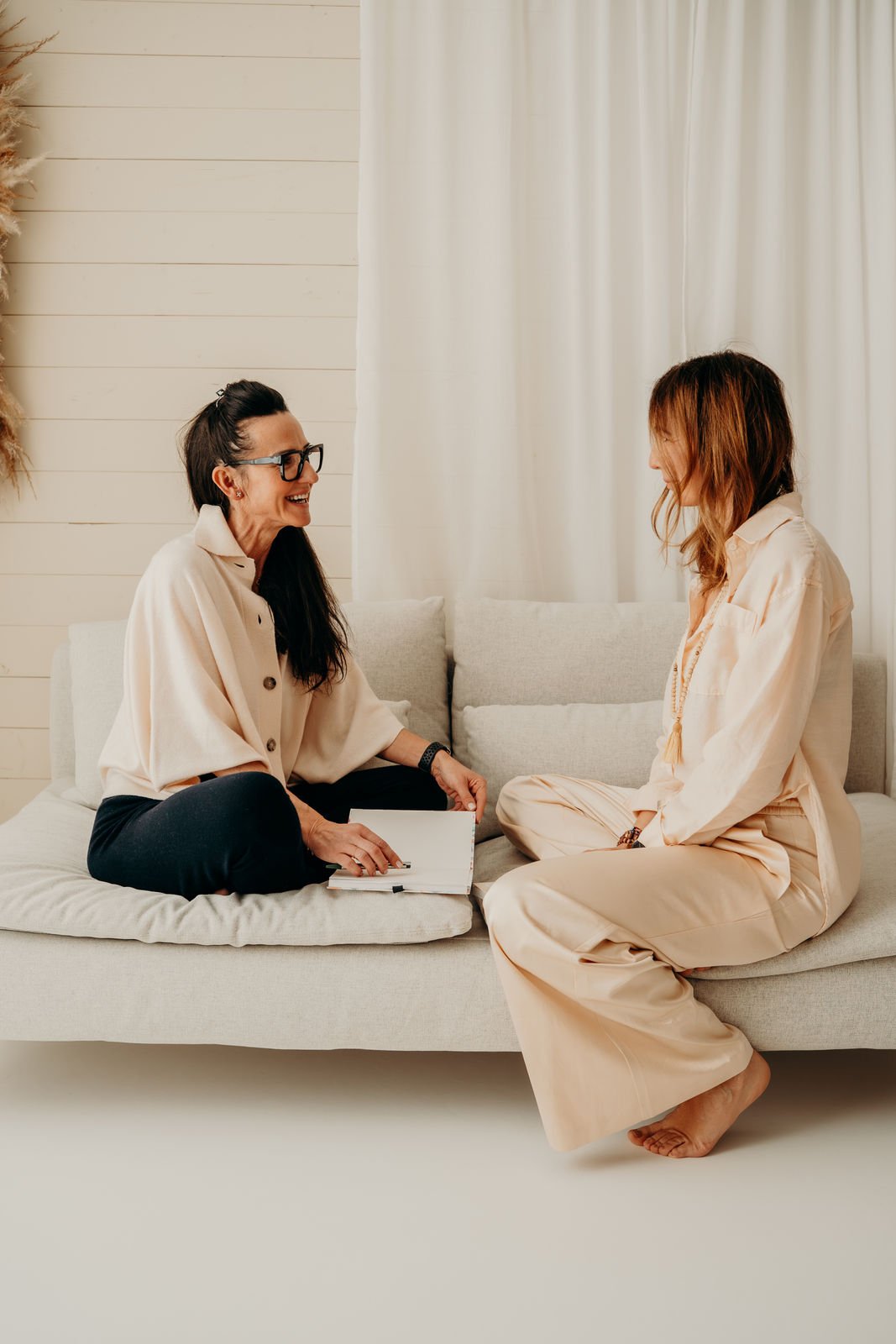 Zwei Frauen sitzen sich auf einem Sofa gegenüber und unterhalten sich. Die eine Frau trägt eine helle Bluse und dunkle Hosen, die andere trägt helle lockere Kleidung. Die Szene findet in einem hellen Raum mit weißen Vorhängen und Holzpaneelen statt.