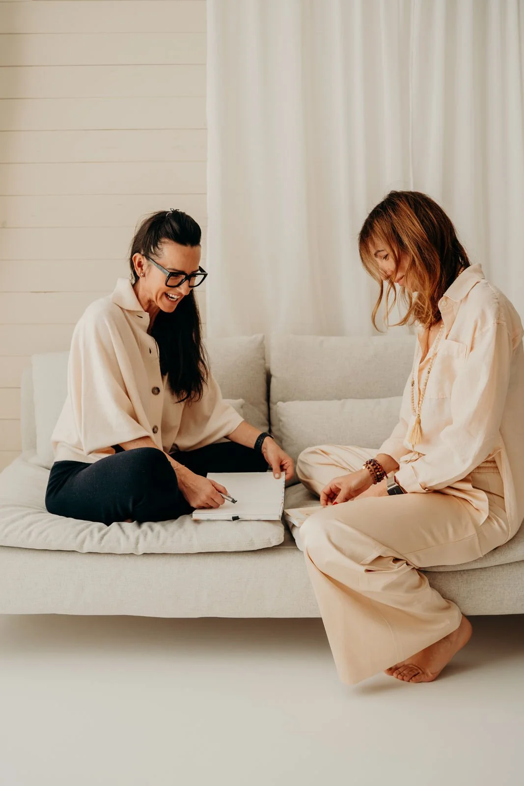 Zwei Frauen sitzen auf einem hellen Sofa, lachen und unterhalten sich. Eine Frau hält einen Notizblock und Stift, die andere hört zu.