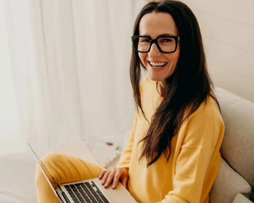 Junge Frau mit dunklen Haaren, Brille und gelbem Outfit sitzt lachend mit Laptop auf einem Bett in einem hellen Raum.