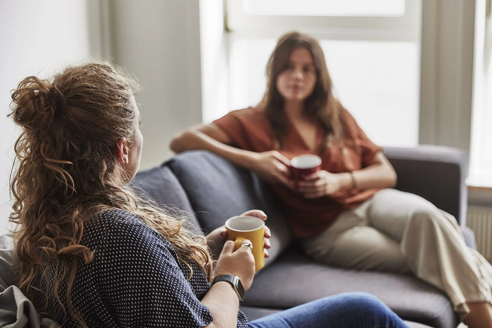 Two women sitting on a couch, having a conversation while holding cups, in a well-lit room with a large window in the background.