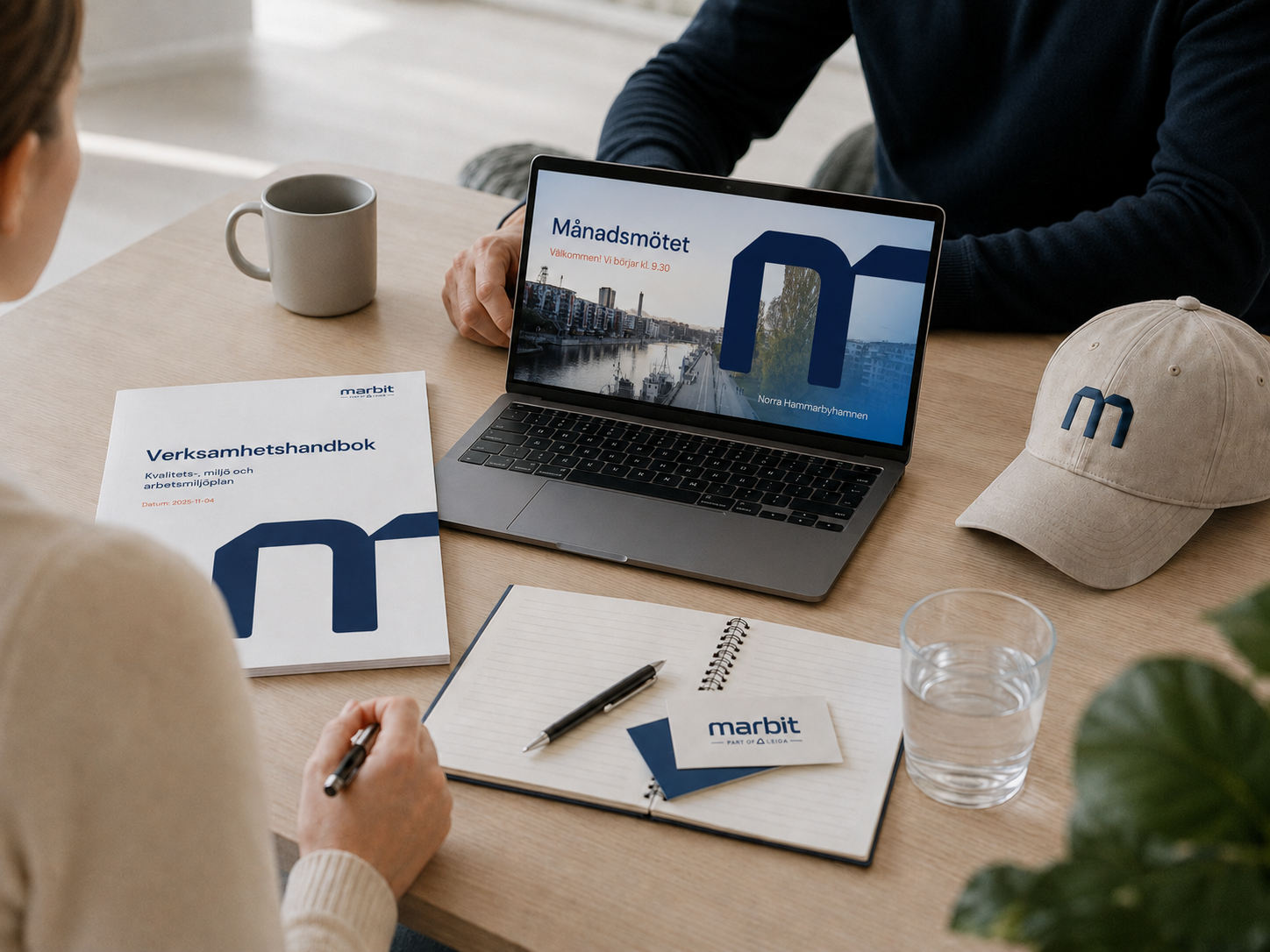 A meeting scene with a woman and a man at a wooden table discussing documents, a laptop displaying a meeting invite, a book titled 'Verksamhetshandbok,' a baseball cap with an 'm' logo, a glass of water, a notepad with a pen, and a coffee mug.