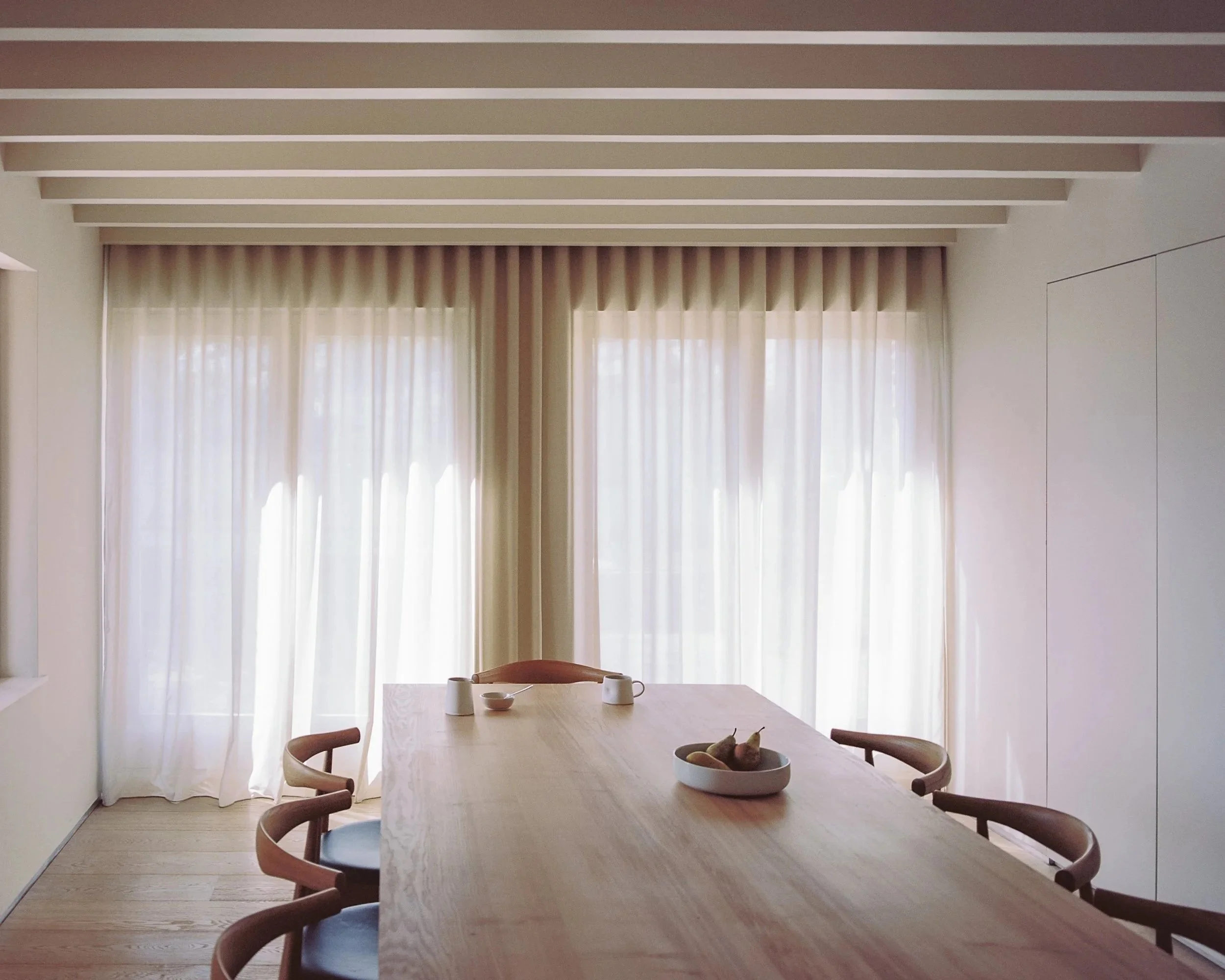 A dining room with a wooden table, six chairs, and a large window with light-colored curtains.