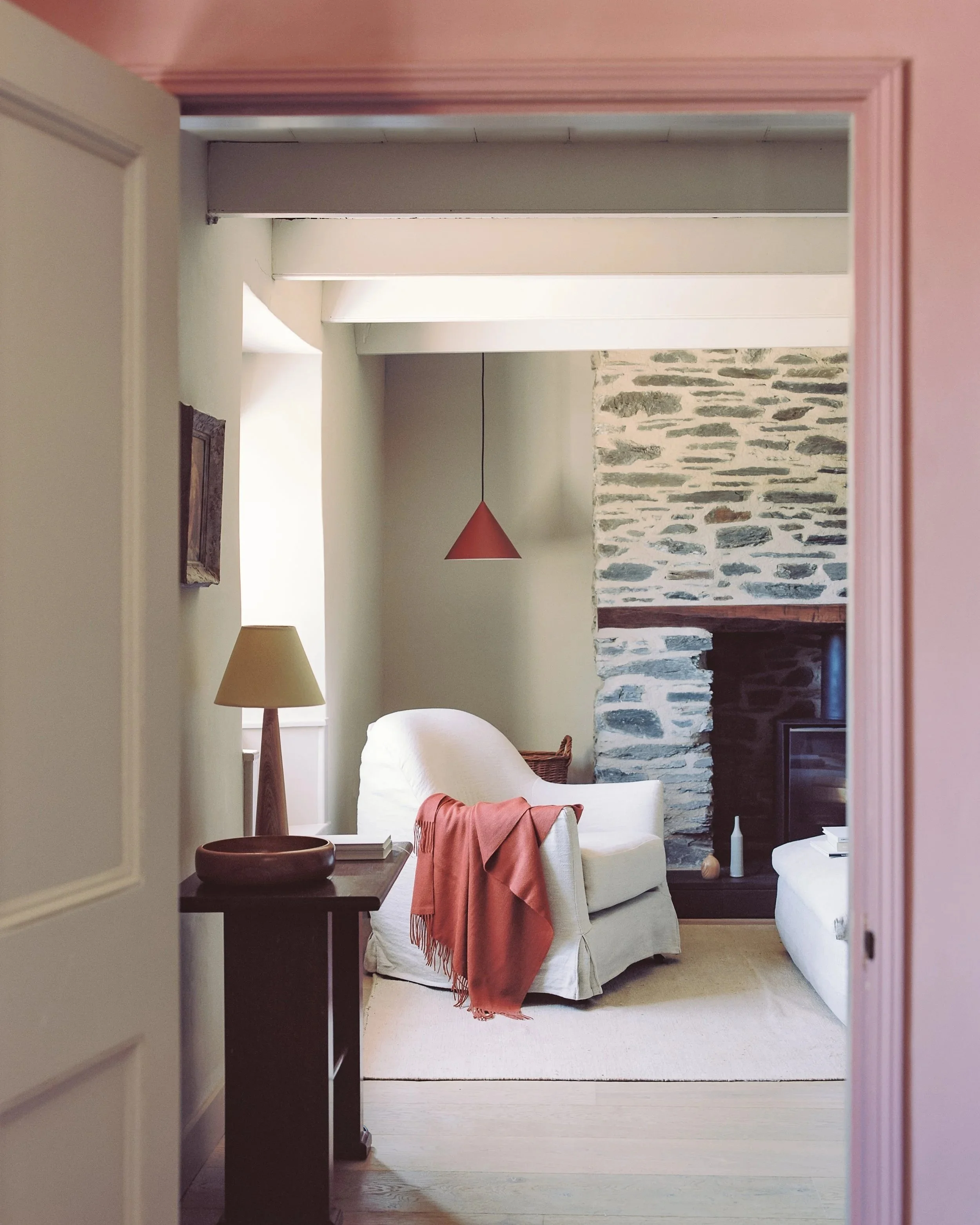 Interior of a living room viewed through a doorway. The room features a stone accent wall, a white armchair with an orange throw blanket, a side table with a lamp, and a red pendant light hanging from the ceiling. There is a flat-screen TV partially 