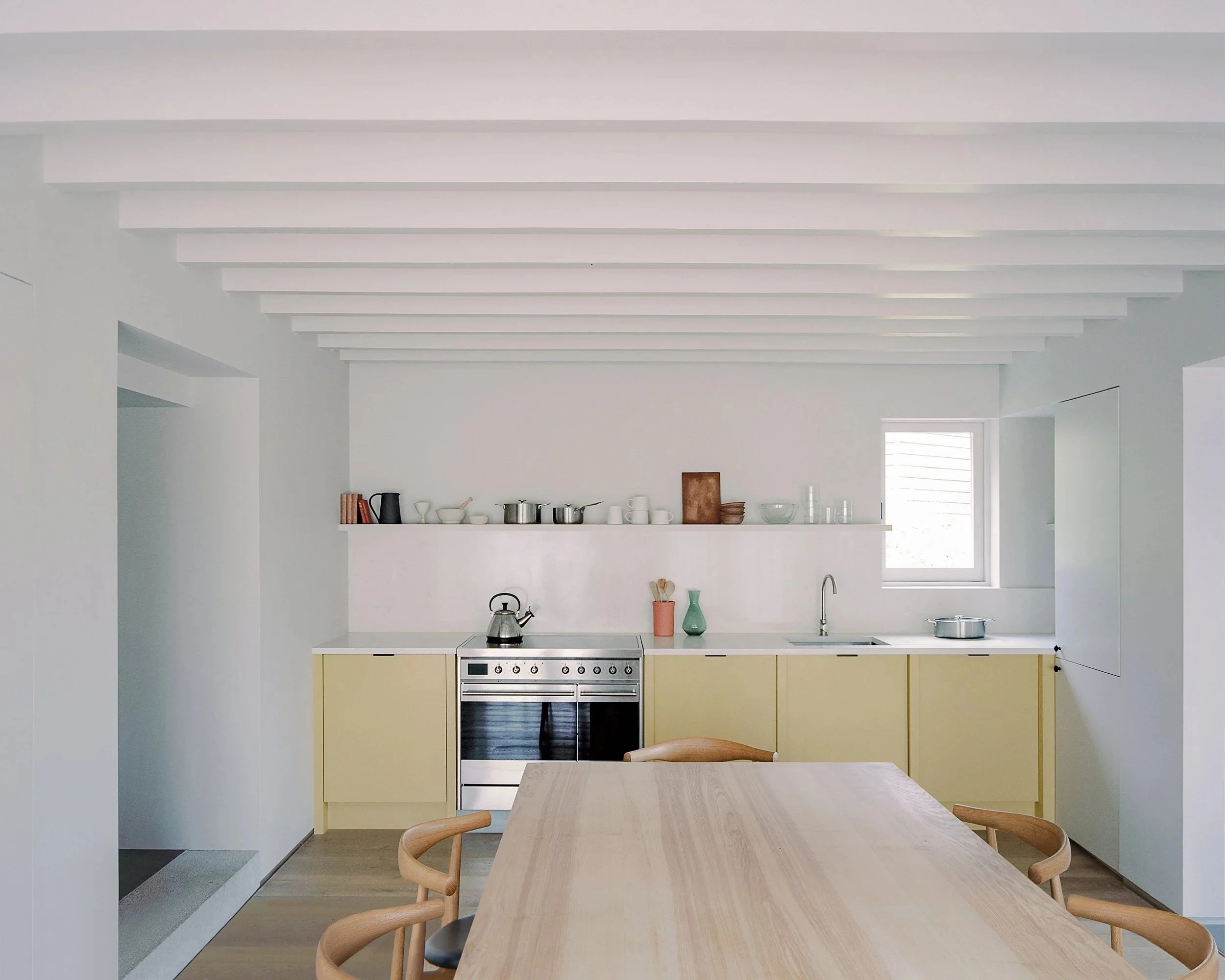 Modern kitchen with white walls and ceiling beams, yellow cabinets, stainless steel oven, and a wooden dining table with chairs.