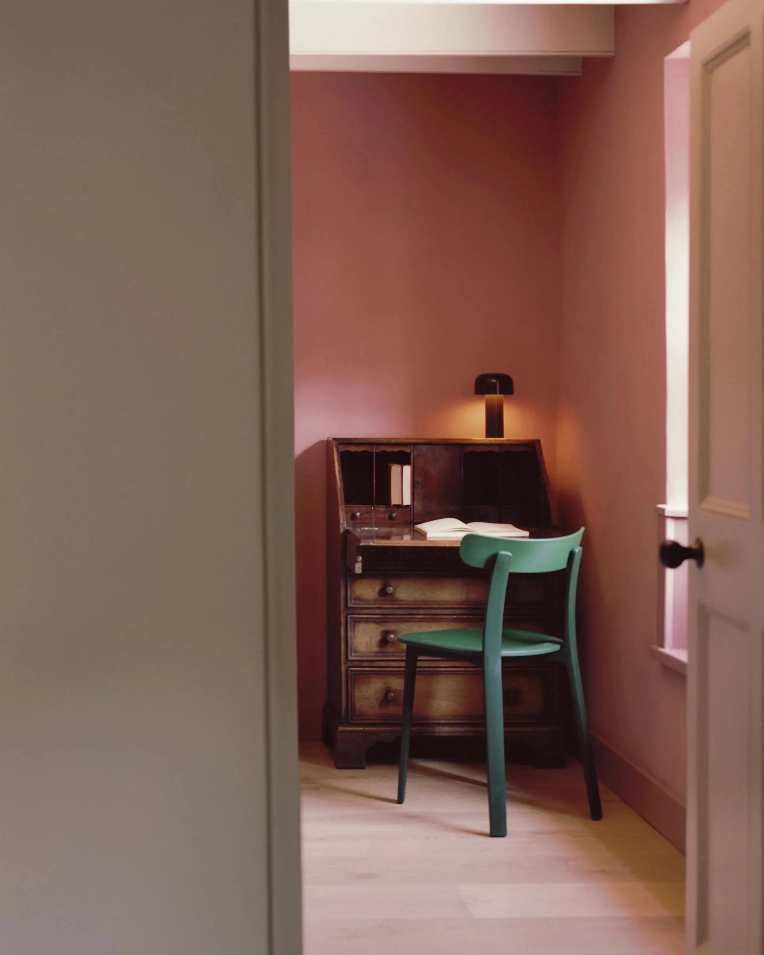 Looking into a room with pink walls, a wooden desk, a green wooden chair, a lamp on the desk, an open book, and a door in the right corner.