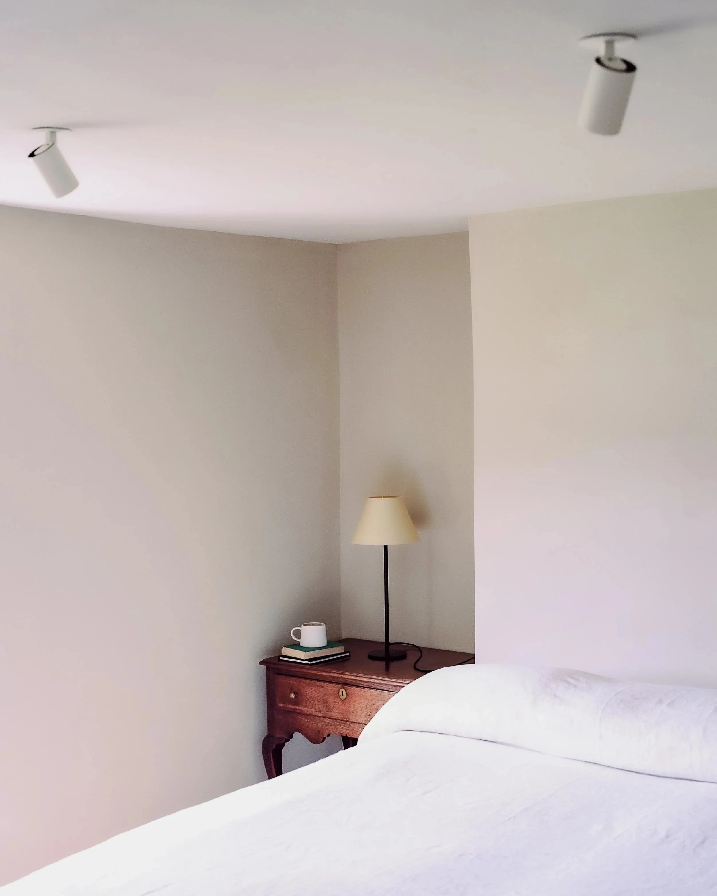 A bedroom corner with a wooden nightstand, a white bed, a table lamp, a white mug, and a couple of books, with beige walls and overhead lighting.