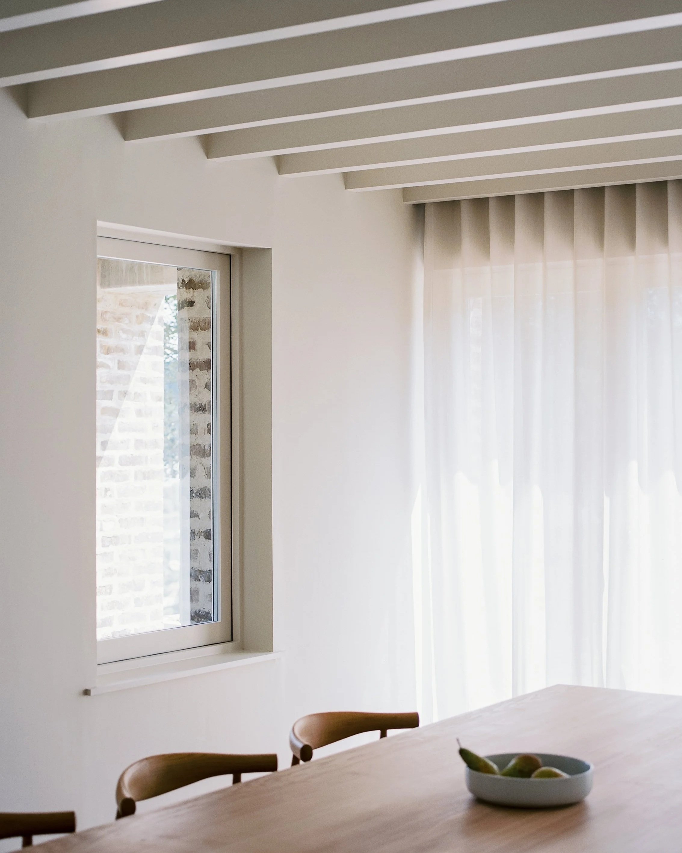 A view of a bright room with a large window, sheer curtains, a wooden table with chairs, and a bowl of fruit in the foreground.