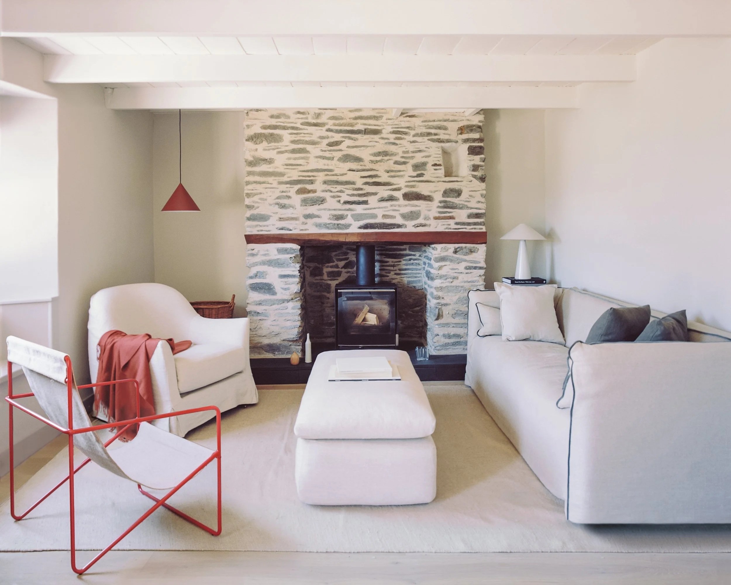 Living room with a stone fireplace, white walls, and a beige carpet. There is a white armchair with a rust-colored blanket, a wicker basket, a white sofa with cushions, a white floor lamp, a small black side table, and a red metal chair with a white 