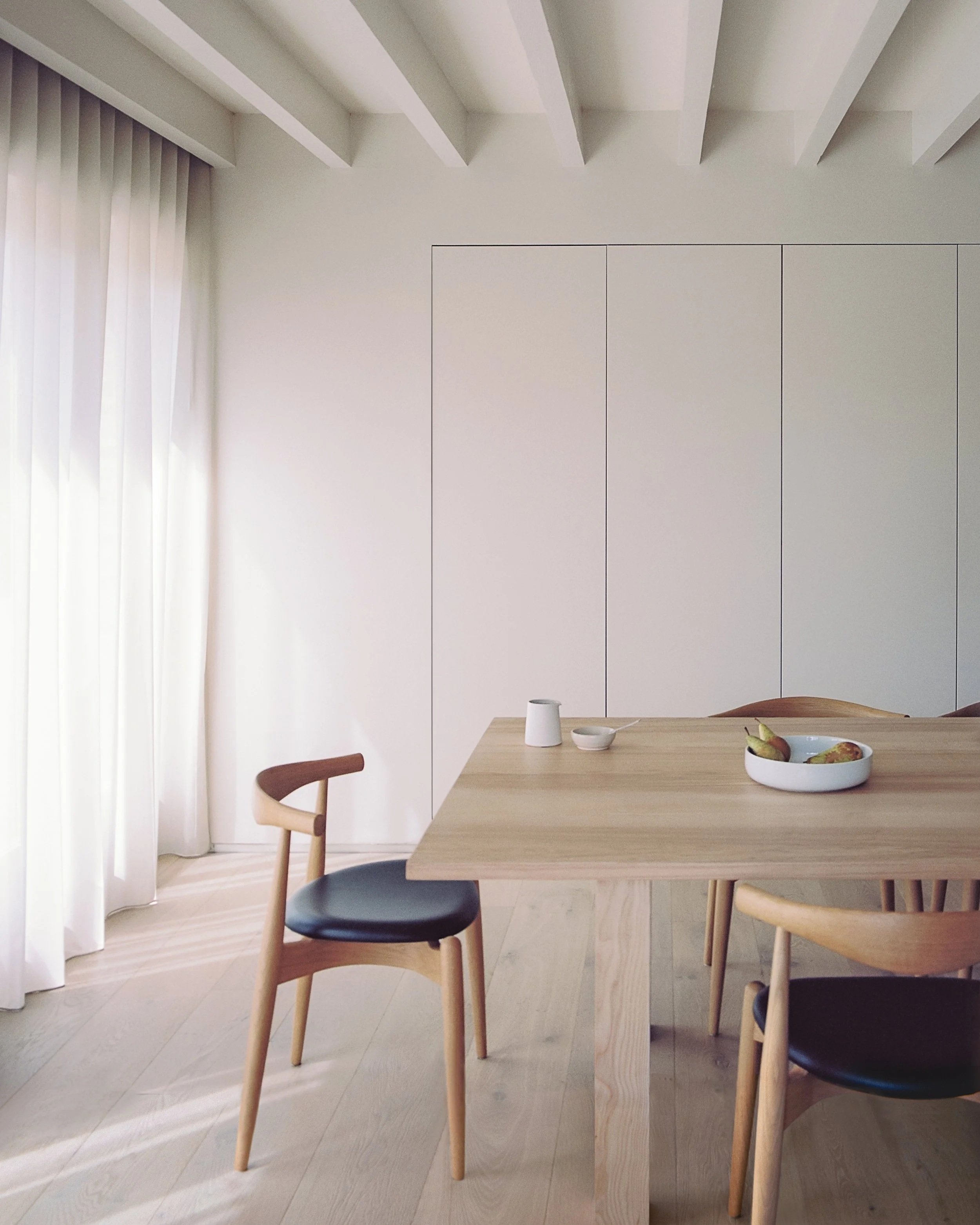 A minimalist dining room with light wooden table, chairs with black and light wood finishes, white wall panels, and sheer white curtains along the window, with a bowl of pears on the table.