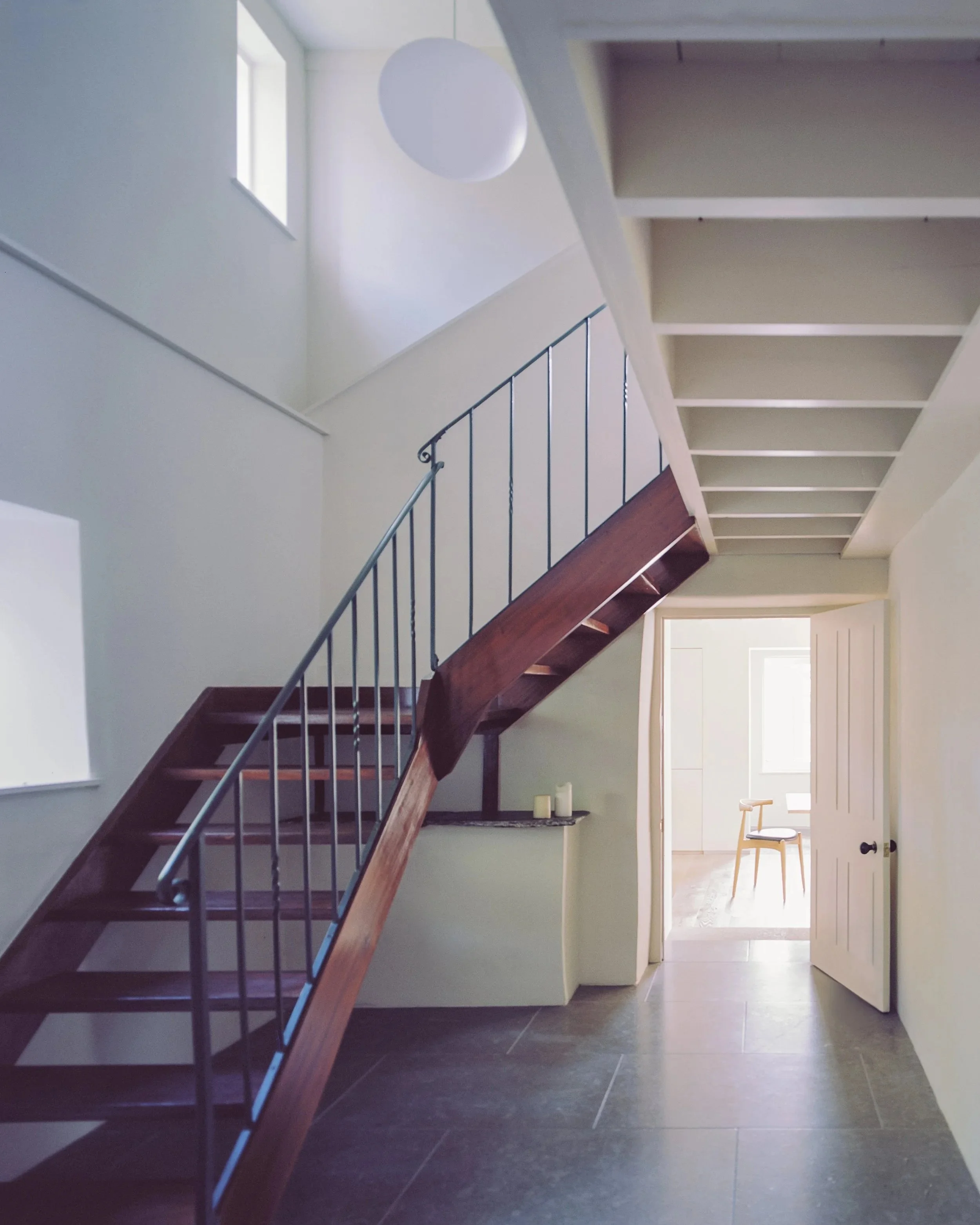 Interior view of a staircase with dark wooden steps and a black metal railing, leading up to a bright landing with natural light coming from a window. An open door reveals a room with a chair and candles on a shelf.