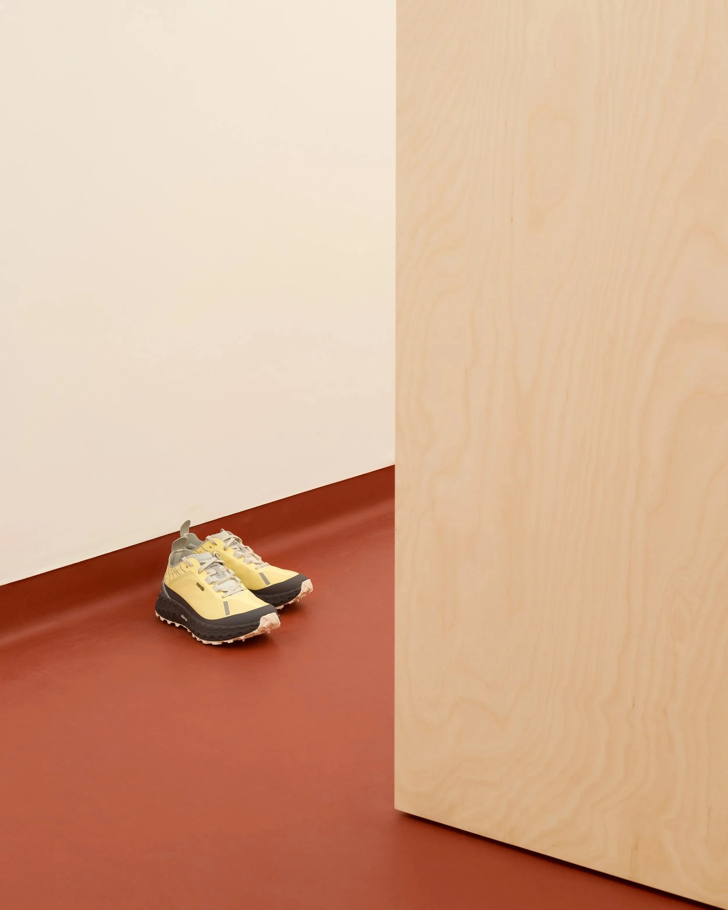 Yellow and gray athletic shoes on a reddish-brown floor next to a light-colored wooden structure or wall.