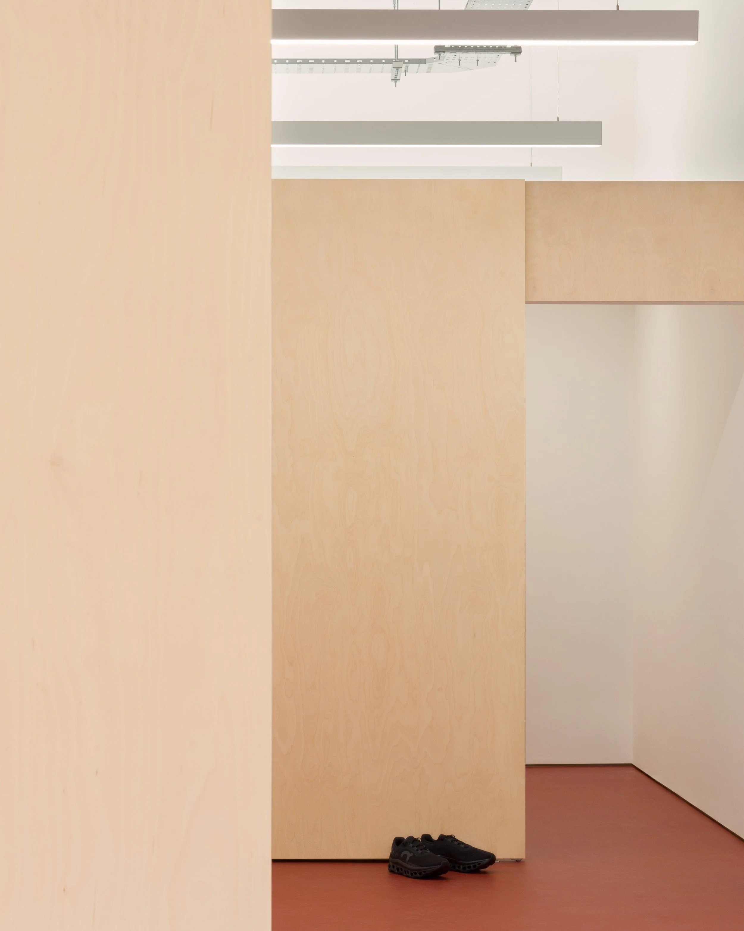 A pair of black shoes on a brown floor near beige wooden panels, with bright white ceiling lights and ceiling ventilation in the background.