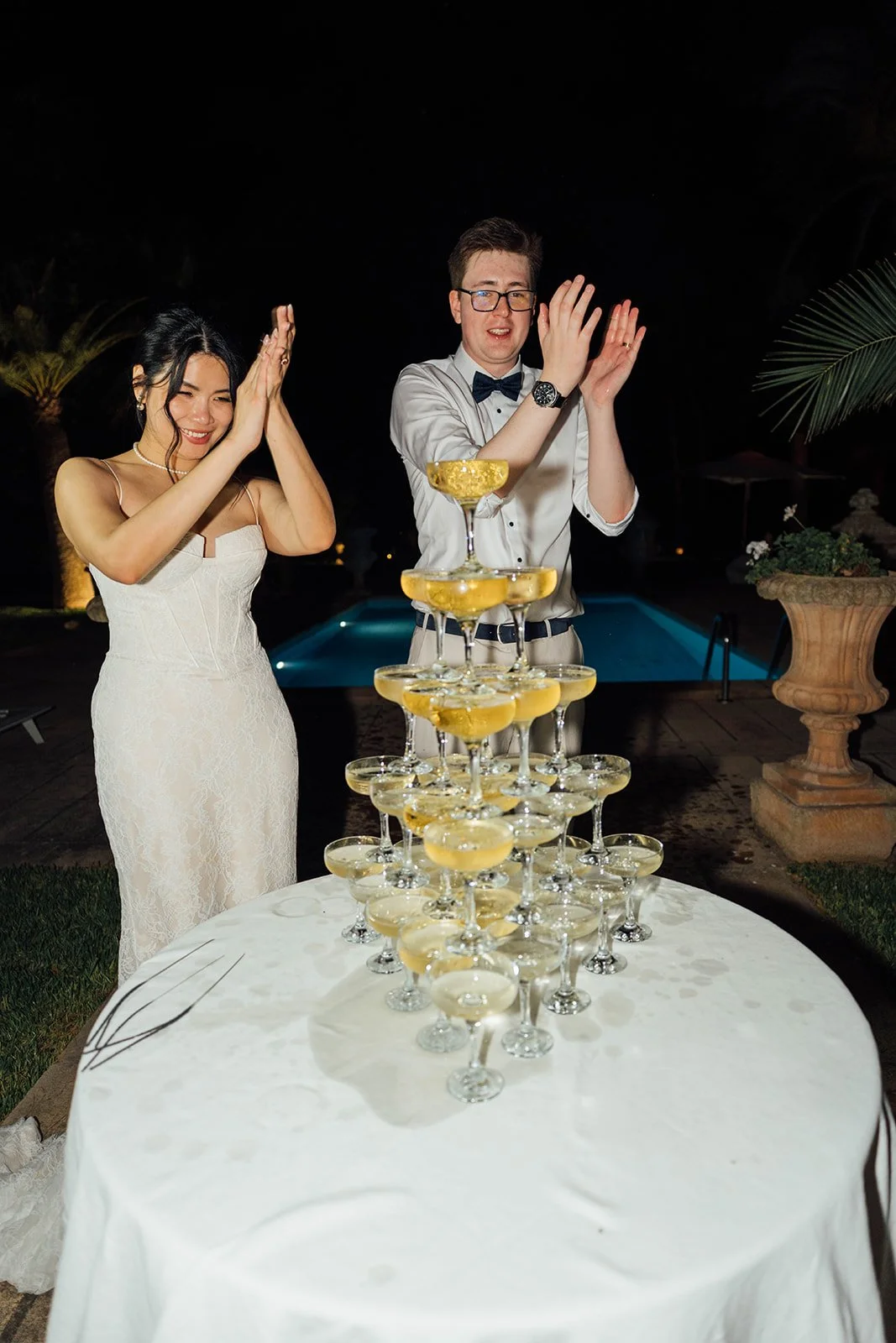 Ein Mann und eine Frau feiern bei Nacht mit einem Champagner-Kuchenvulkan, in einem Garten mit Pool und Palmen, beide in formeller Kleidung, lächelnd, applaudierend.