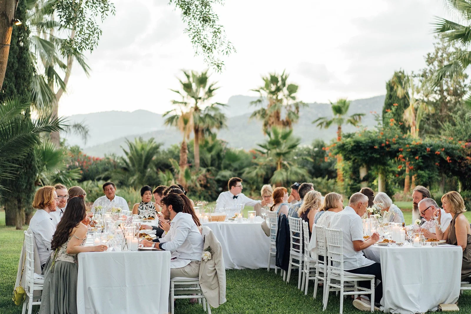 Eine große Gruppe von Menschen sitzt an einem langen, weißen Esstisch im Freien, umgeben von Palmen und üppiger Vegetation, bei einem festlichen Abendessen im Sonnenschein.