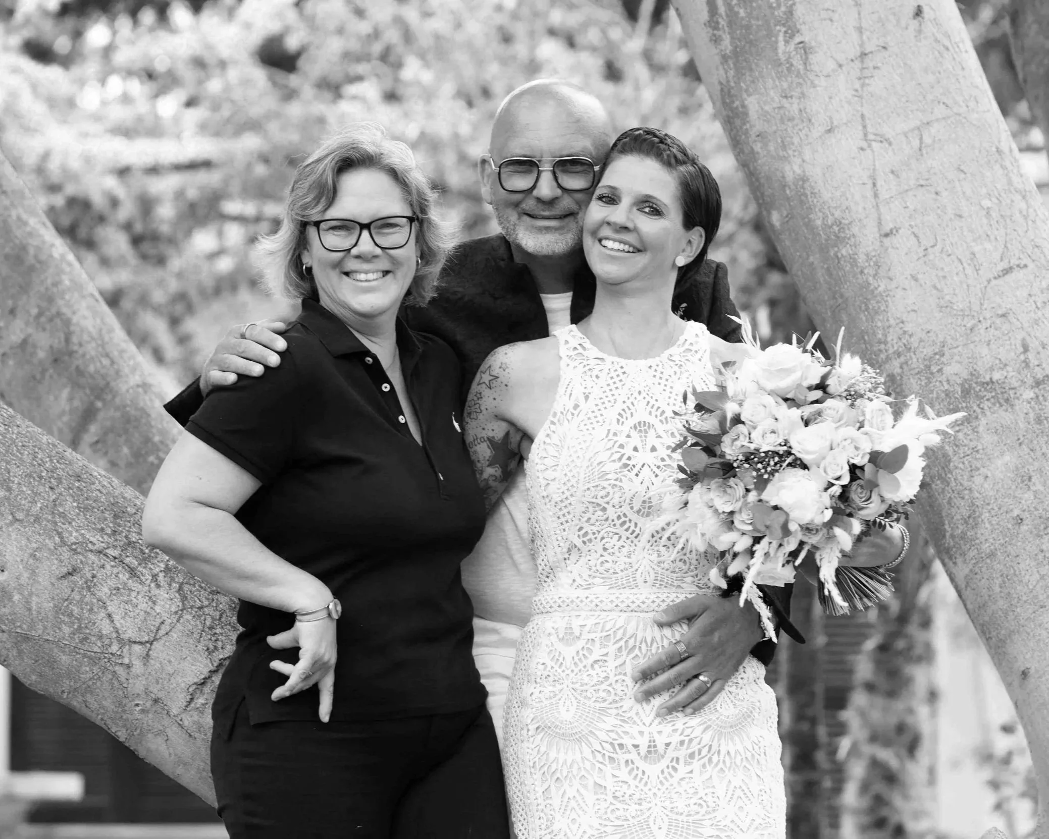 Alexandra mit ihrem Brautpaar, zwei Frauen und ein Mann, bei einer Feier im Freien, lachend, umarmend, mit Blumen. Frau am rechten trägt ein weißes Kleid, Mann in der Mitte trägt Brille, Frau an der linken trägt schwarze Kleidung und Brille.
