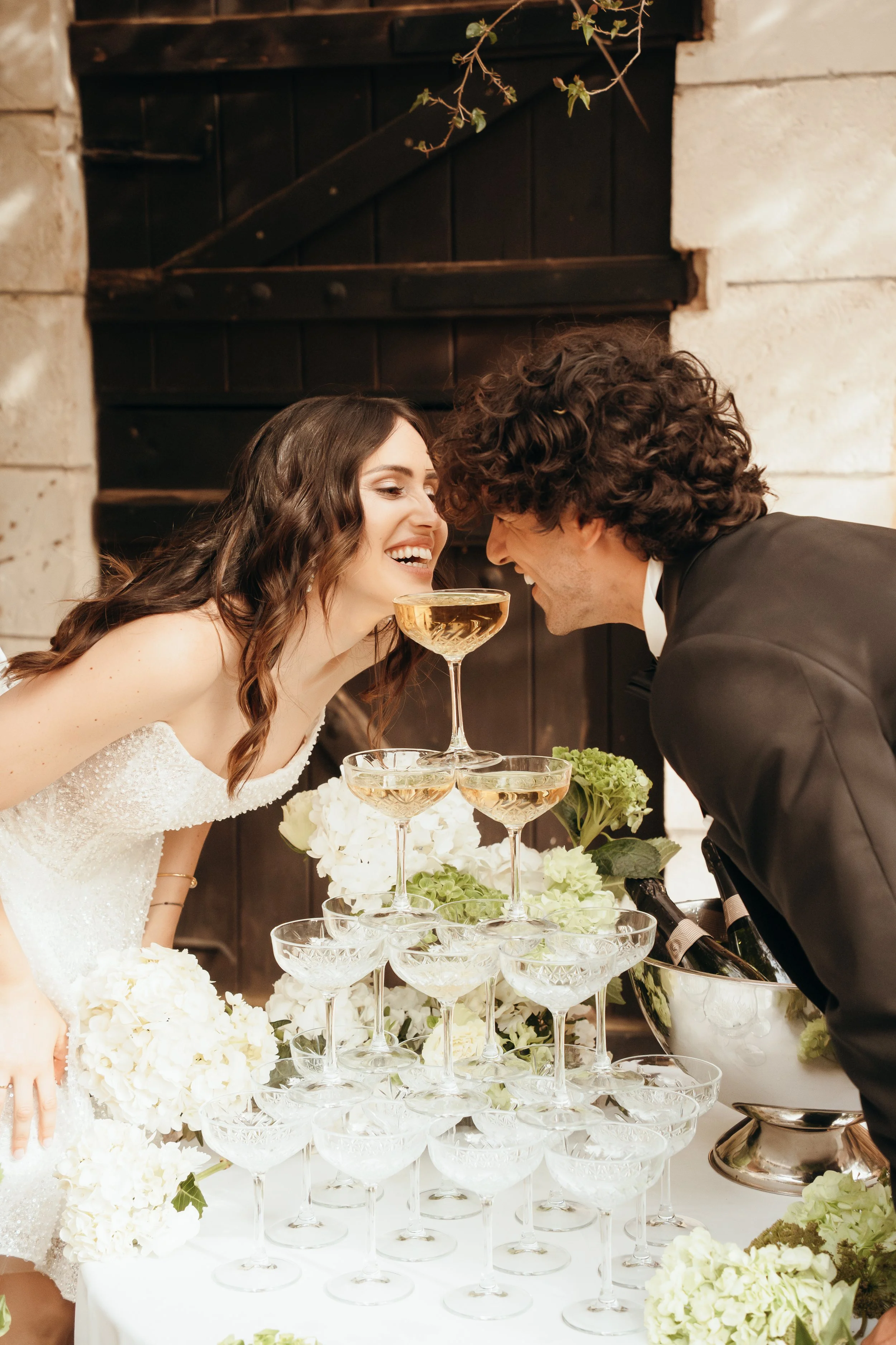 Ein Hochzeitspaar stoßt mit Champagnergläsern an, während sie sich vor einem Turm aus leeren Champagnergläsern auf einem Tisch im Festsaal begrüßen.