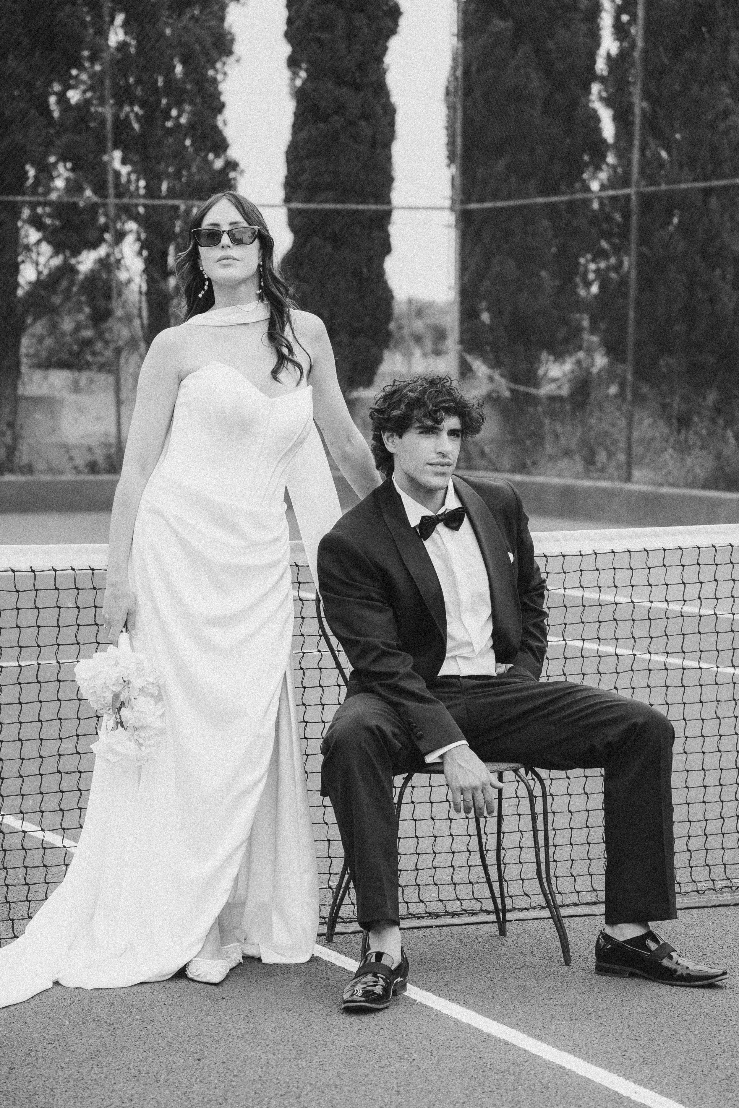 Schwarz-weiße Fotografie eines Paares auf einem Tennisplatz. Die Frau trägt ein elegantes Hochzeitskleid, sunglasses und Ohrringe, während der Mann Anzug und Fliege trägt, auf einem Stuhl sitzend.