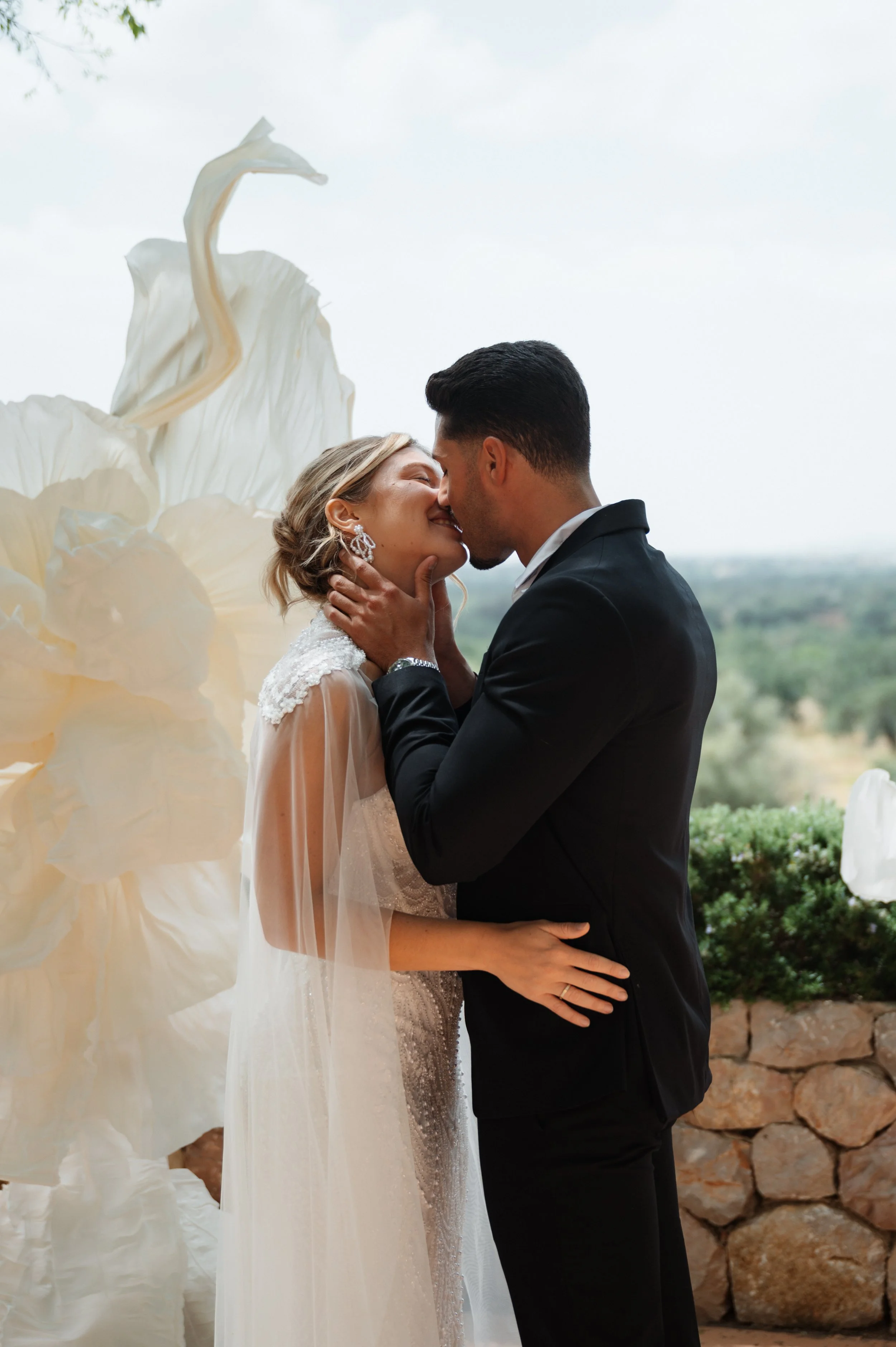 Ein Brautpaar bei einer Hochzeit, die sich küssen, im Freien vor 2 Giant Flowers, der neuesten Deko für Hochzeiten 2026/27. Die Braut trägt ein weißes Kleid mit Spitze und großen Ohrringen, der Bräutigam trägt einen schwarzen Anzug.