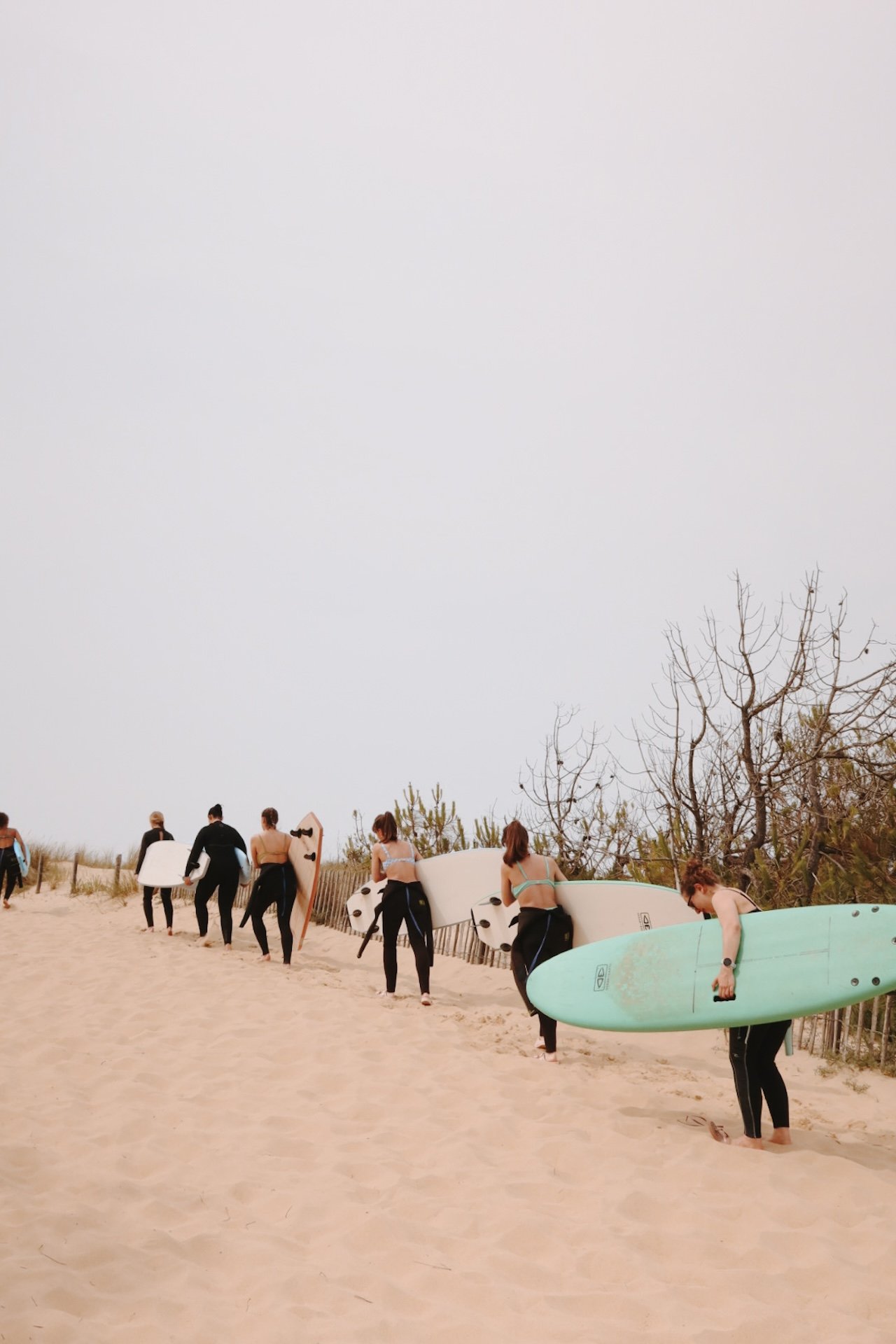 Groupe marchant pieds nus dans les dunes, planche de surf à la main, lumière dorée du soir — esprit d'aventure d'une retraite yoga et surf