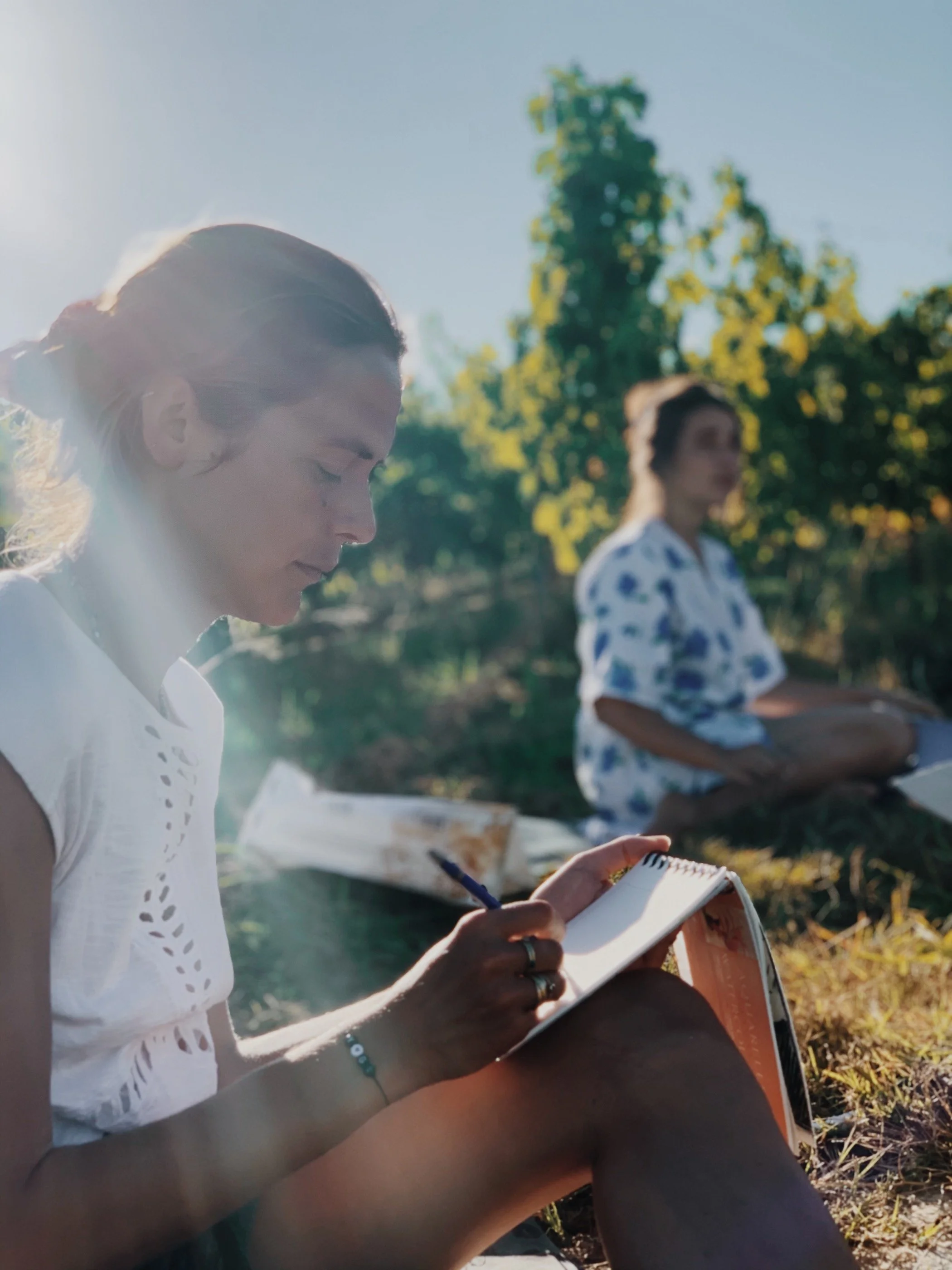 Deux participantes écrivant et dessinant en plein air, traversées par un rayon de lumière — atelier créatif et journaling en pleine nature lors d'une retraite yoga