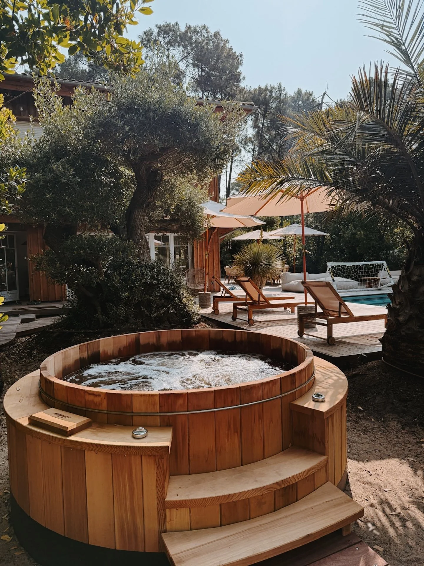 Bain nordique en bois fumant au cœur du jardin, piscine et transats en arrière-plan — détente après les sessions de surf dans une retraite yoga Sud-Ouest